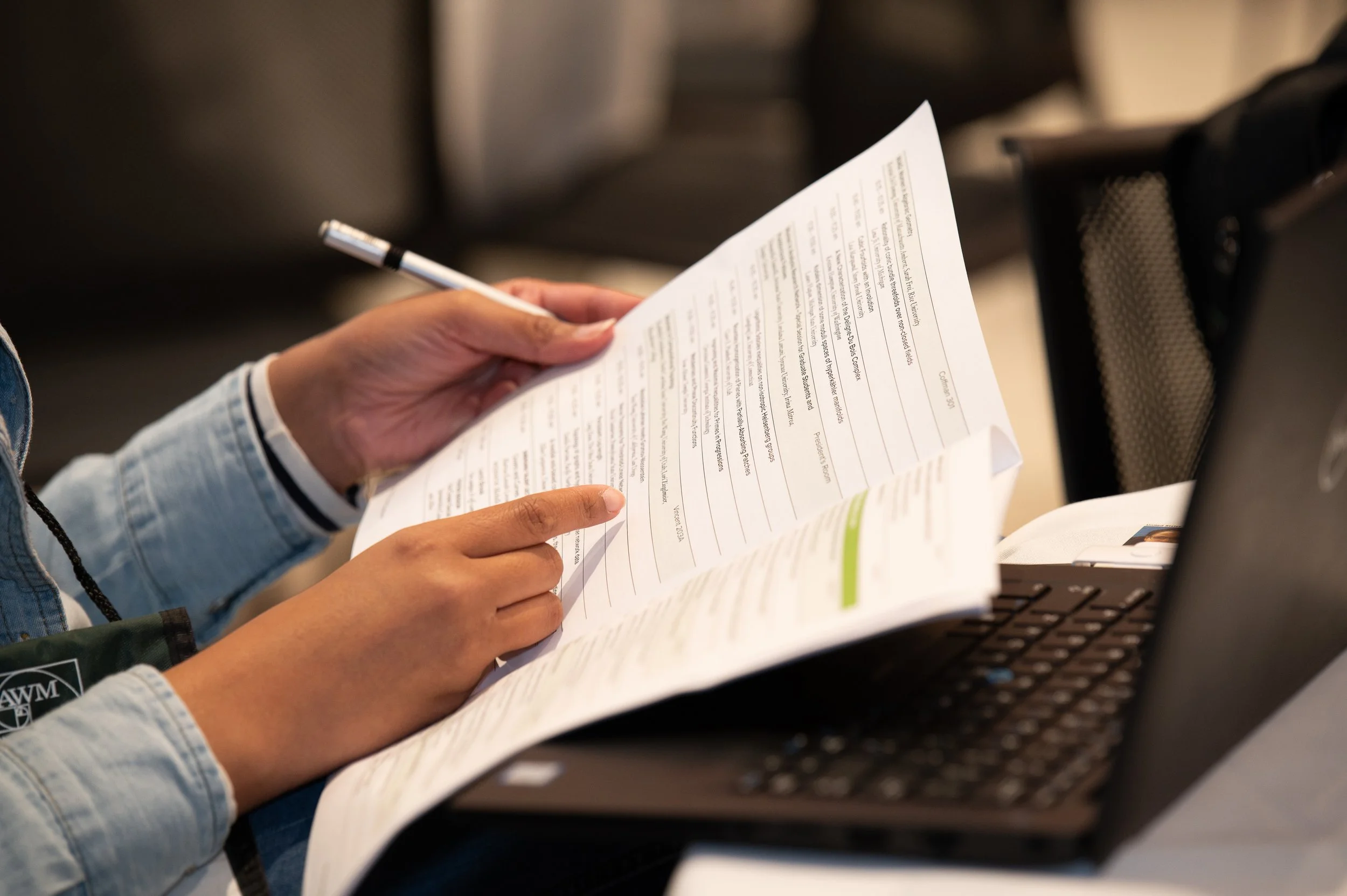 Person holding a white document or booklet with printed text, sitting at a desk with a laptop nearby.