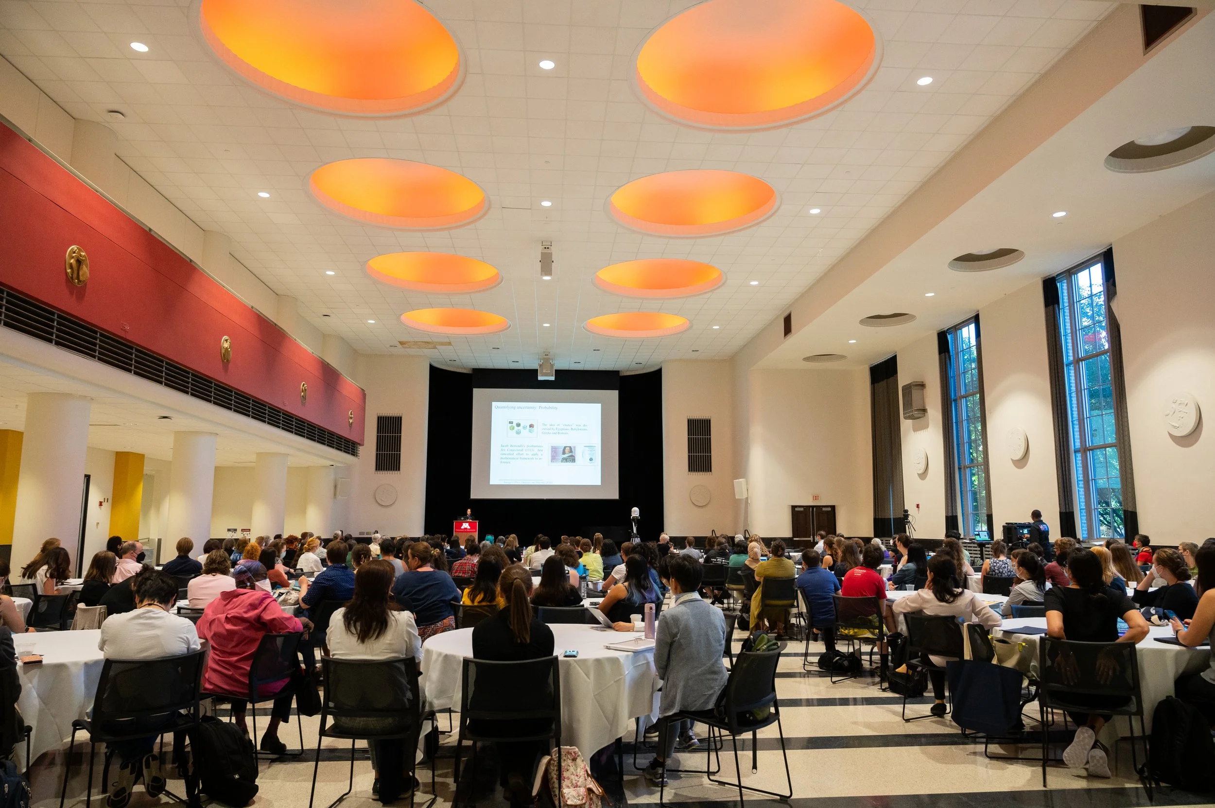 A large conference room filled with people seated at round tables, listening to a presentation. The room has high ceilings with several circular skylights with orange lighting. There is a stage at the front with a large screen displaying a presentati