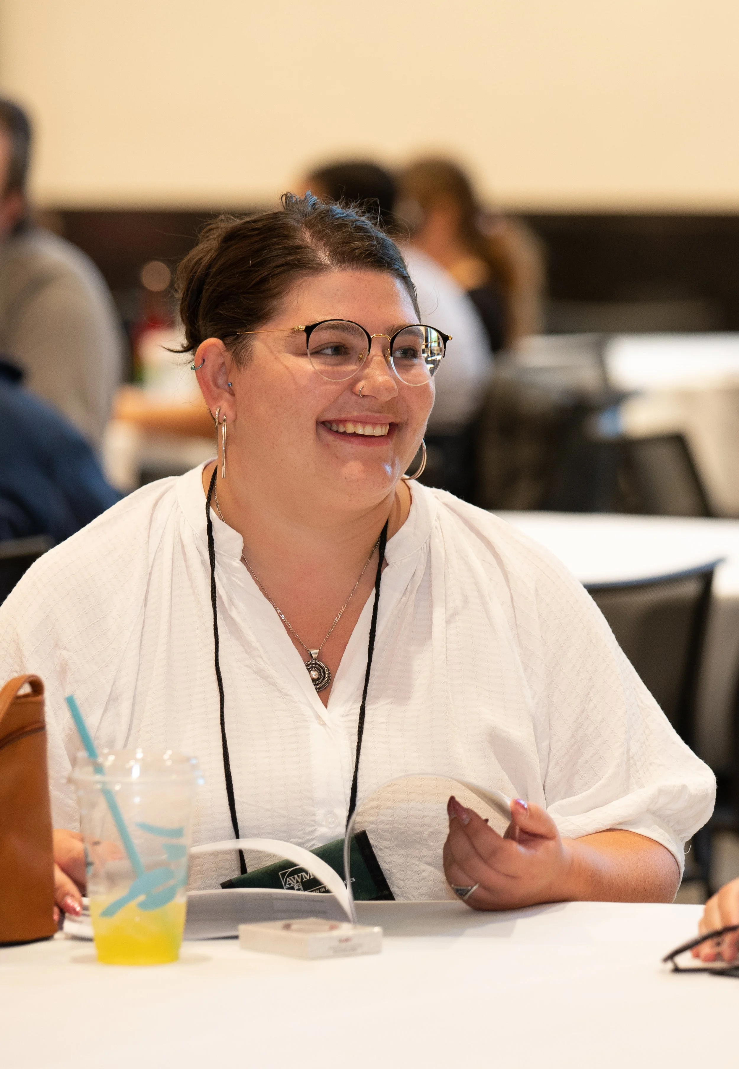 A smiling woman wearing glasses, earrings, and a white top sitting at a table in a group setting, with drinks and papers on the table.