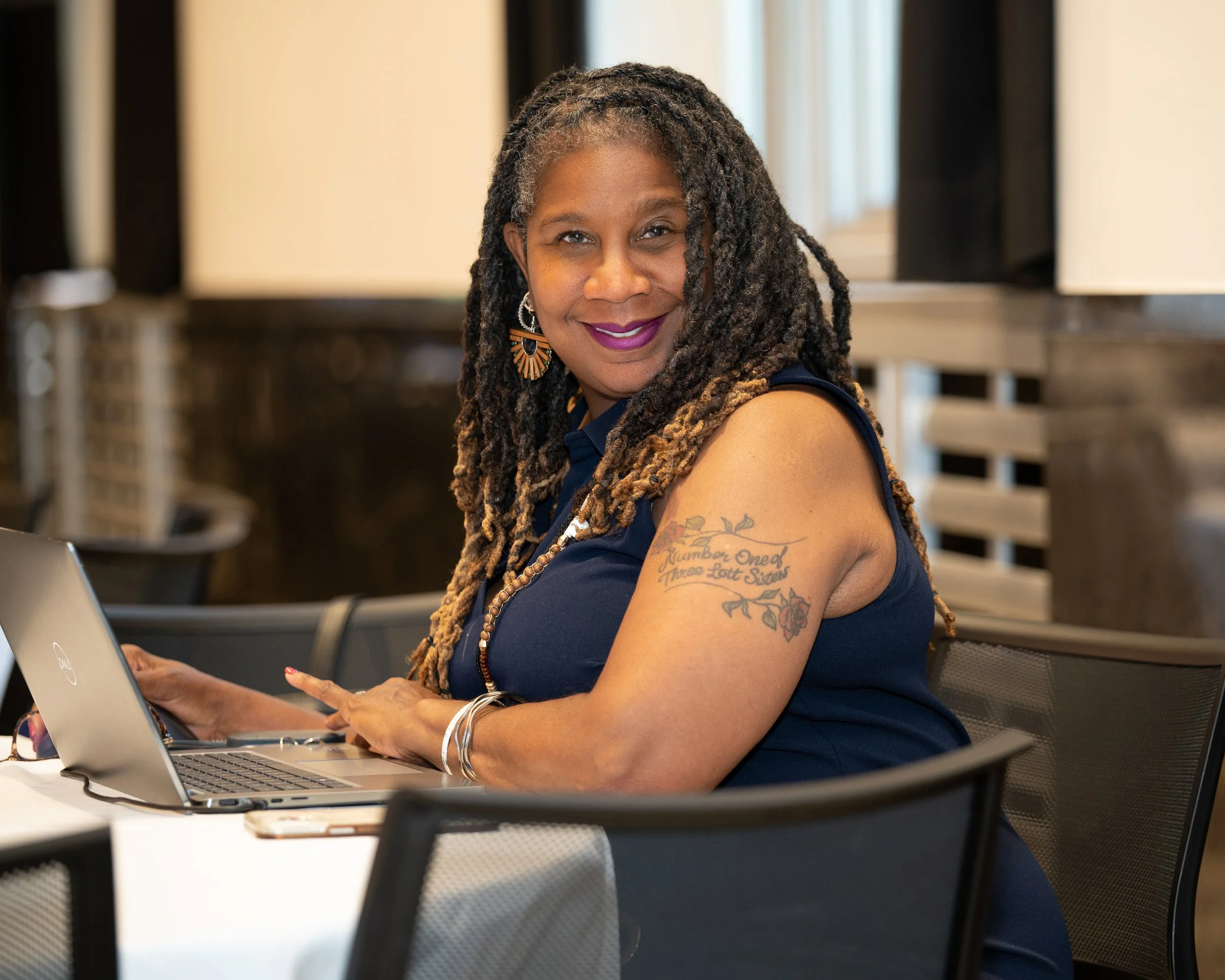 A woman with long dreadlocks, wearing earrings and a sleeveless top, sitting at a table with a laptop, smiling at the camera.