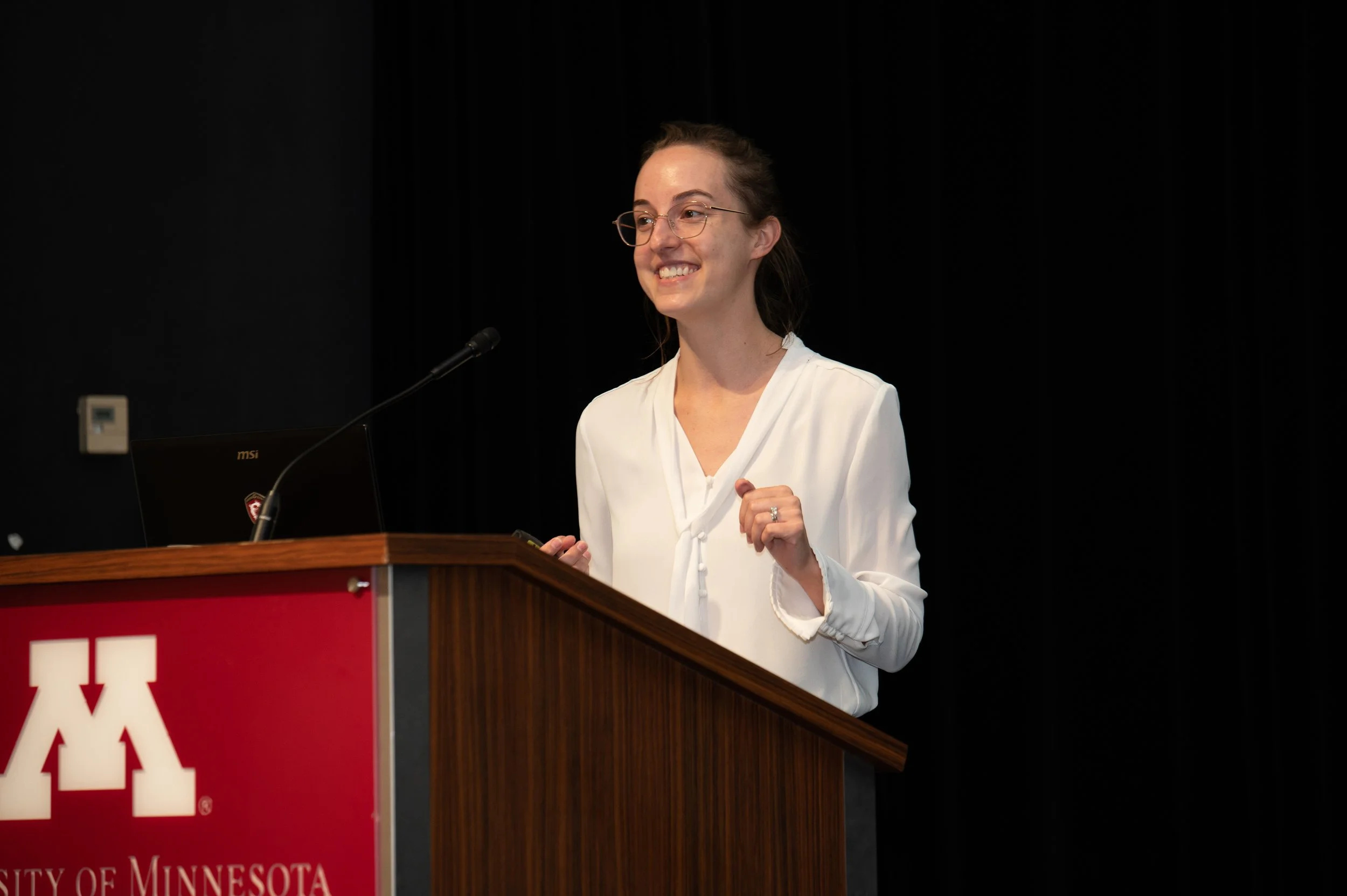 A woman with glasses and a white blouse speaking at a podium with a University of Minnesota sign.