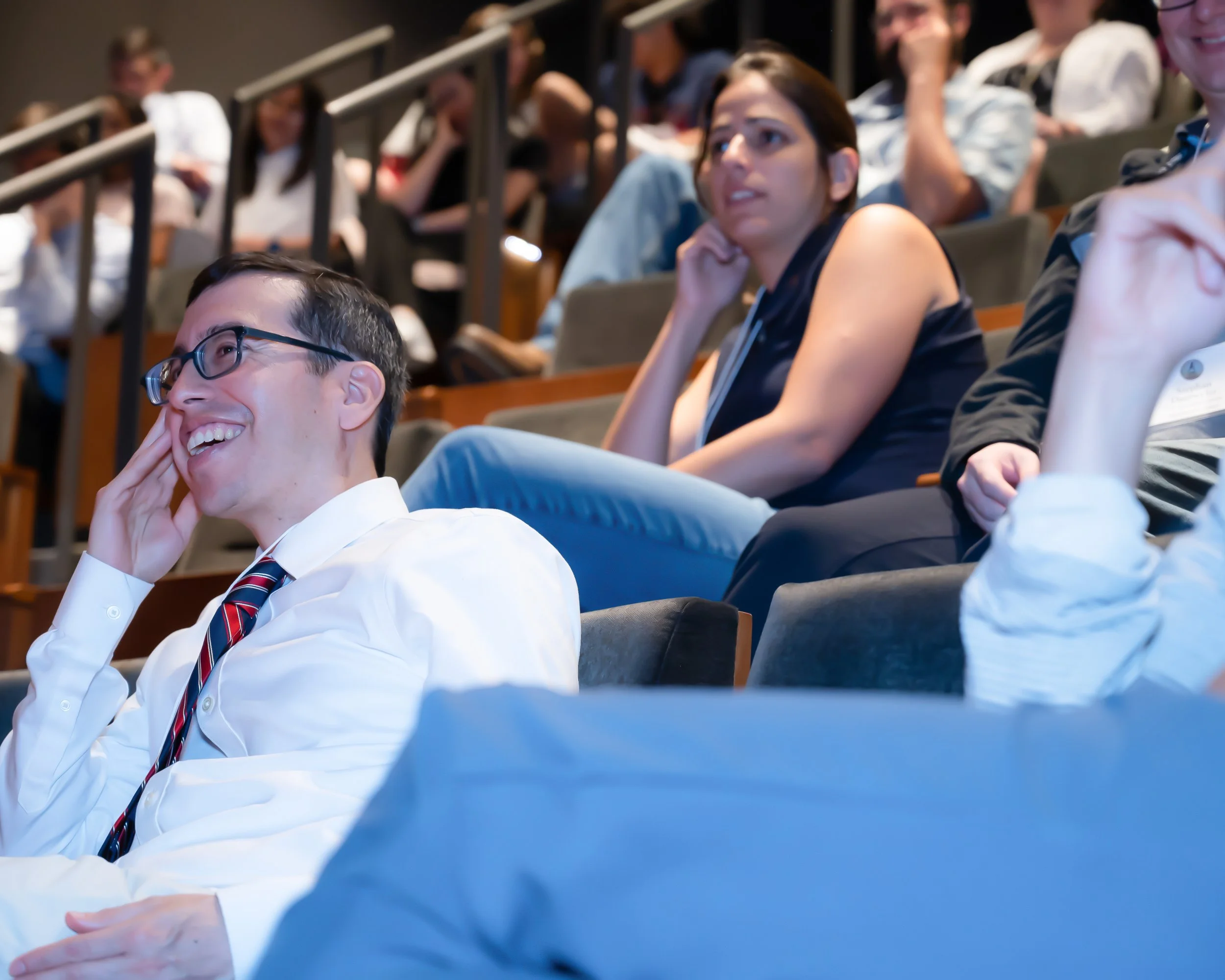 People attending a lecture or conference in a large auditorium, sitting in rows of seats, some smiling and others appearing attentive.
