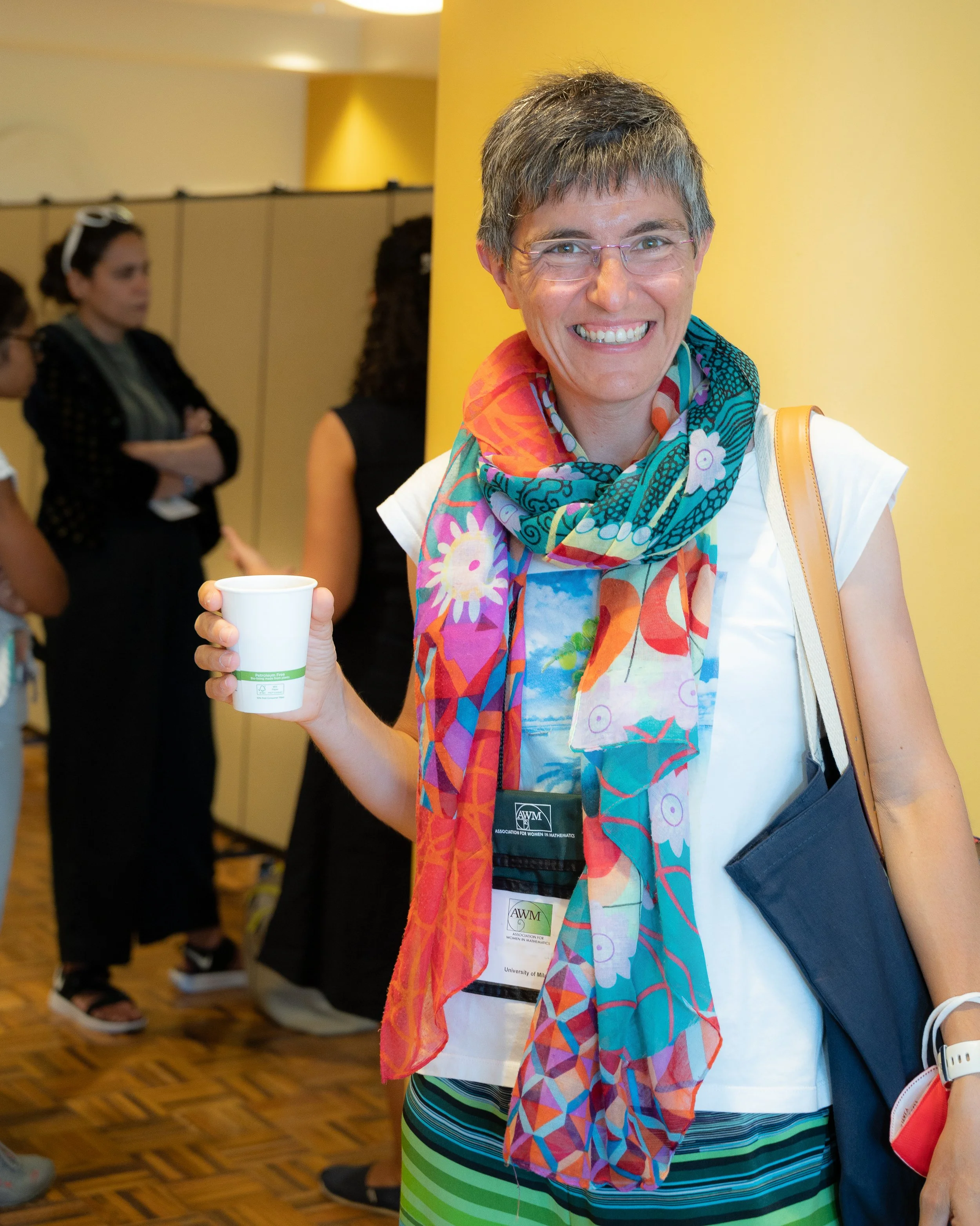 A smiling woman with short brown hair, glasses, and a colorful scarf, holding a white cup, standing in a room with a yellow wall and other people in the background.