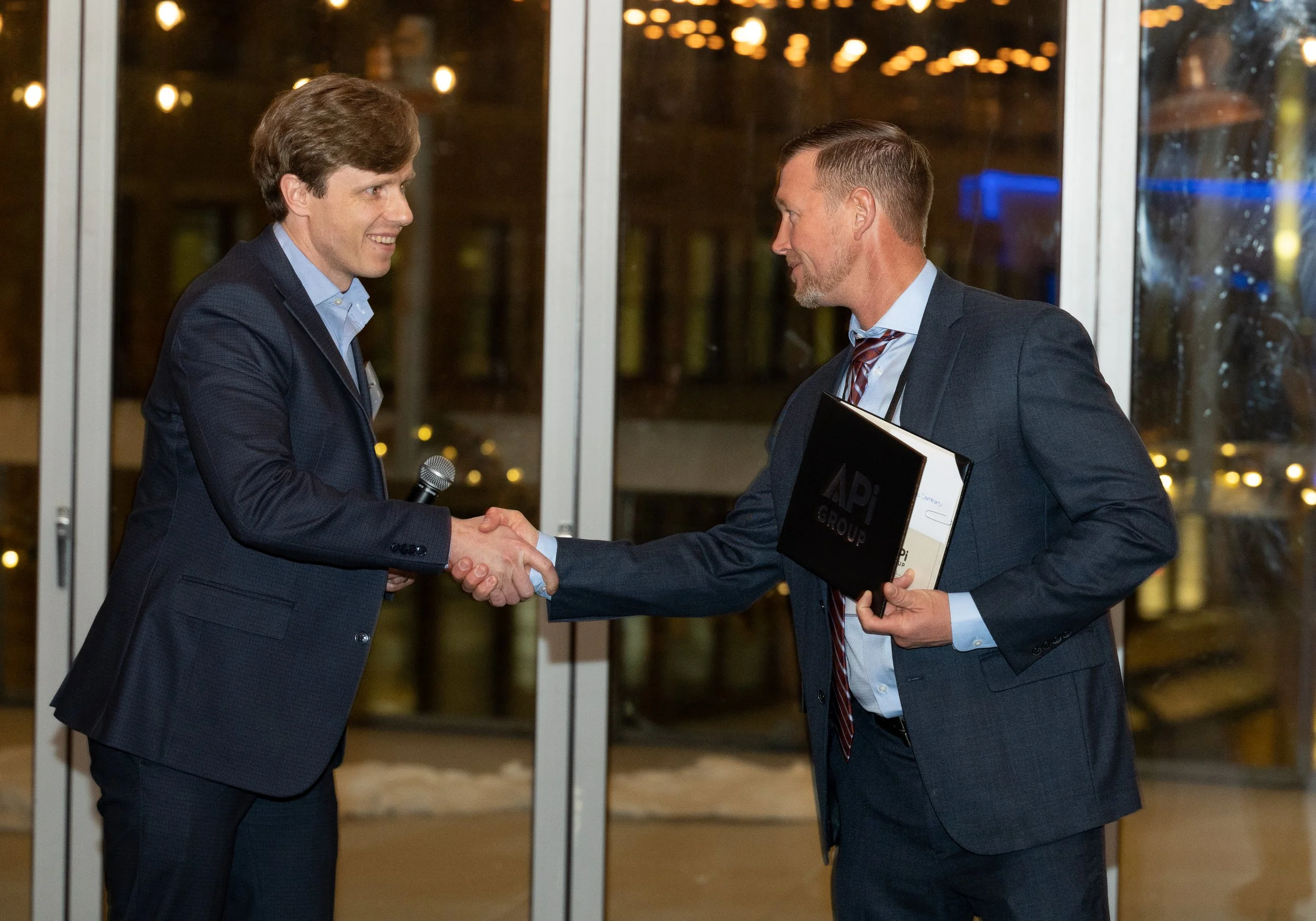 Two men in suits shaking hands at a professional event, one holding a folder and the other a microphone, in front of a glass door with city lights visible outside.