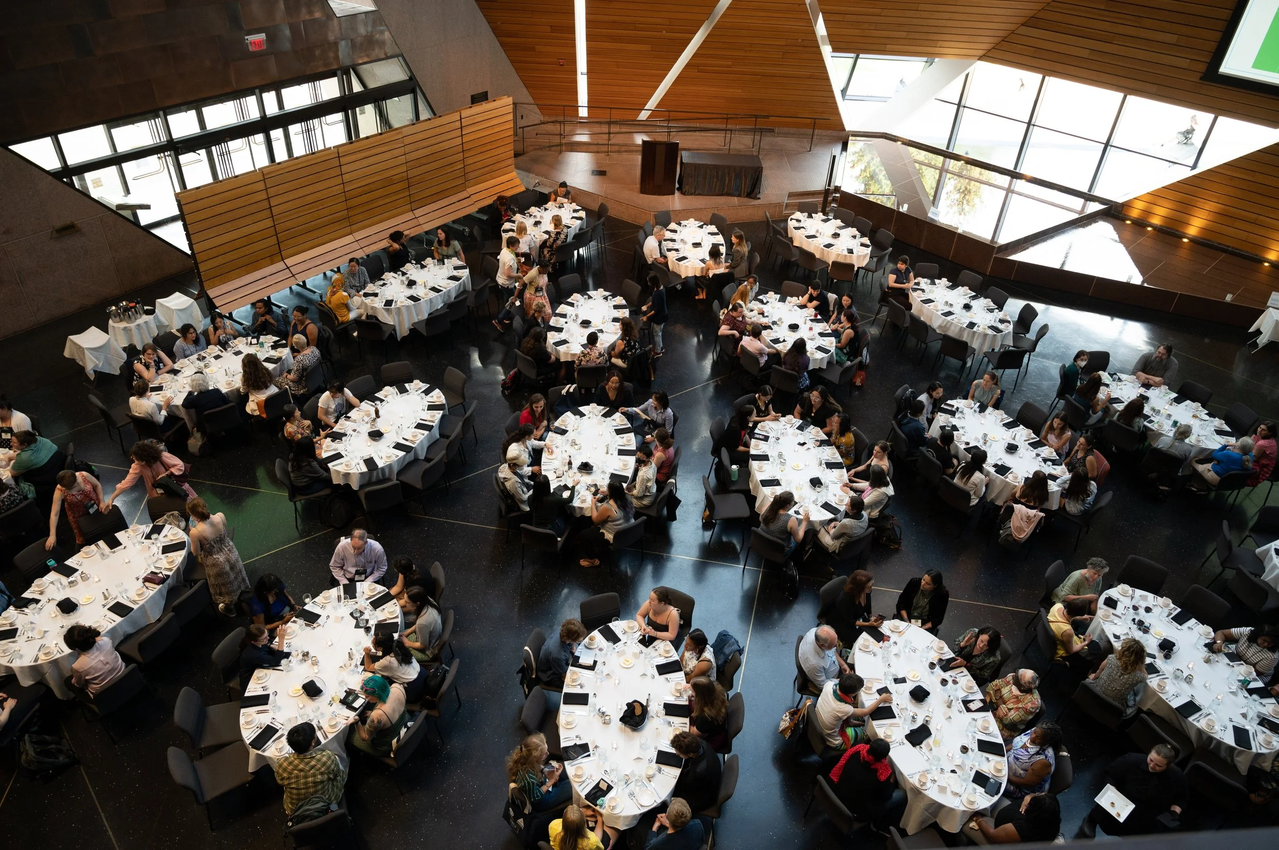 An aerial view of a large banquet hall filled with round tables set for a meal, with many people seated and some walking around, featuring a modern interior with wooden accents and large windows letting in natural light.