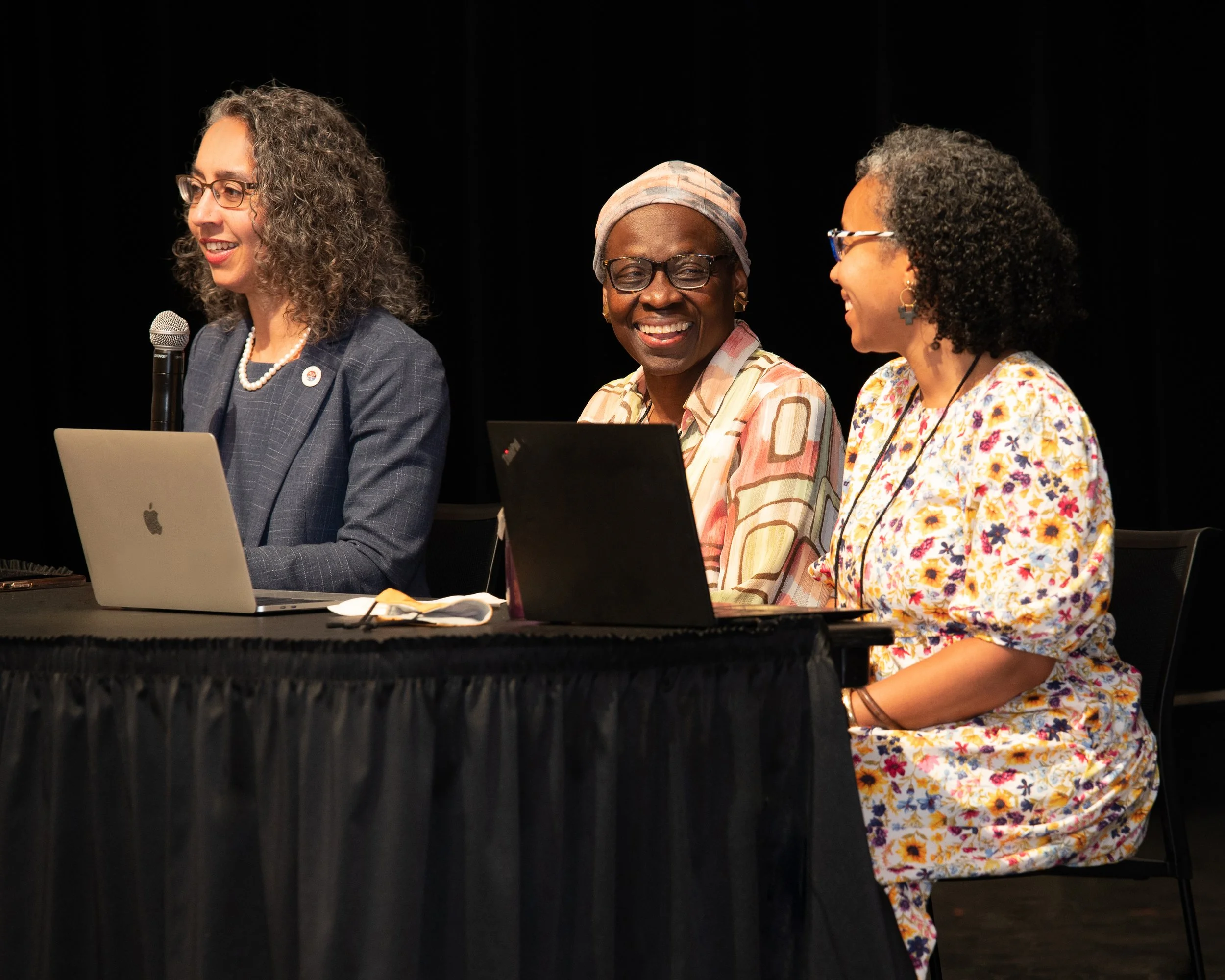 Three women sitting at a table with laptops, engaged in conversation, with a black background.