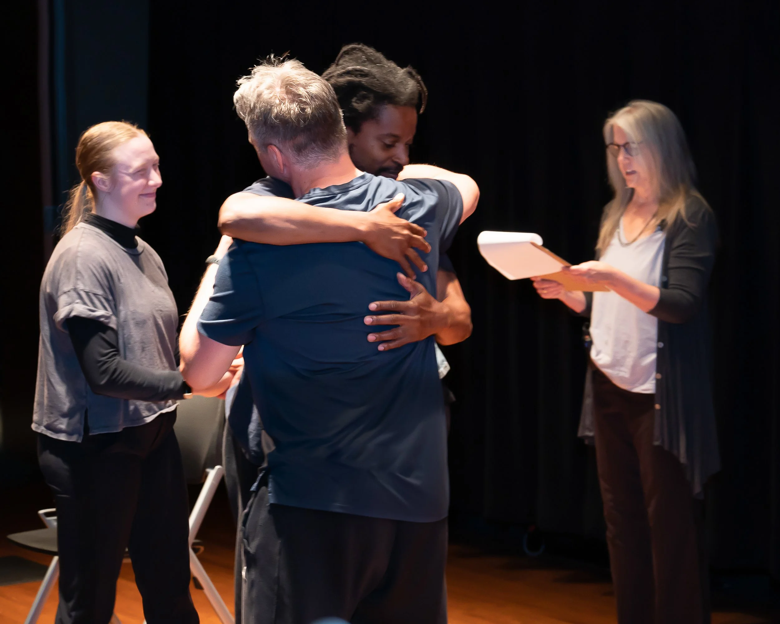 Four people in an emotional moment on a dark stage. Two people are hugging, one smiling woman and another with glasses. Two women are standing nearby, one smiling and one reading from a paper.