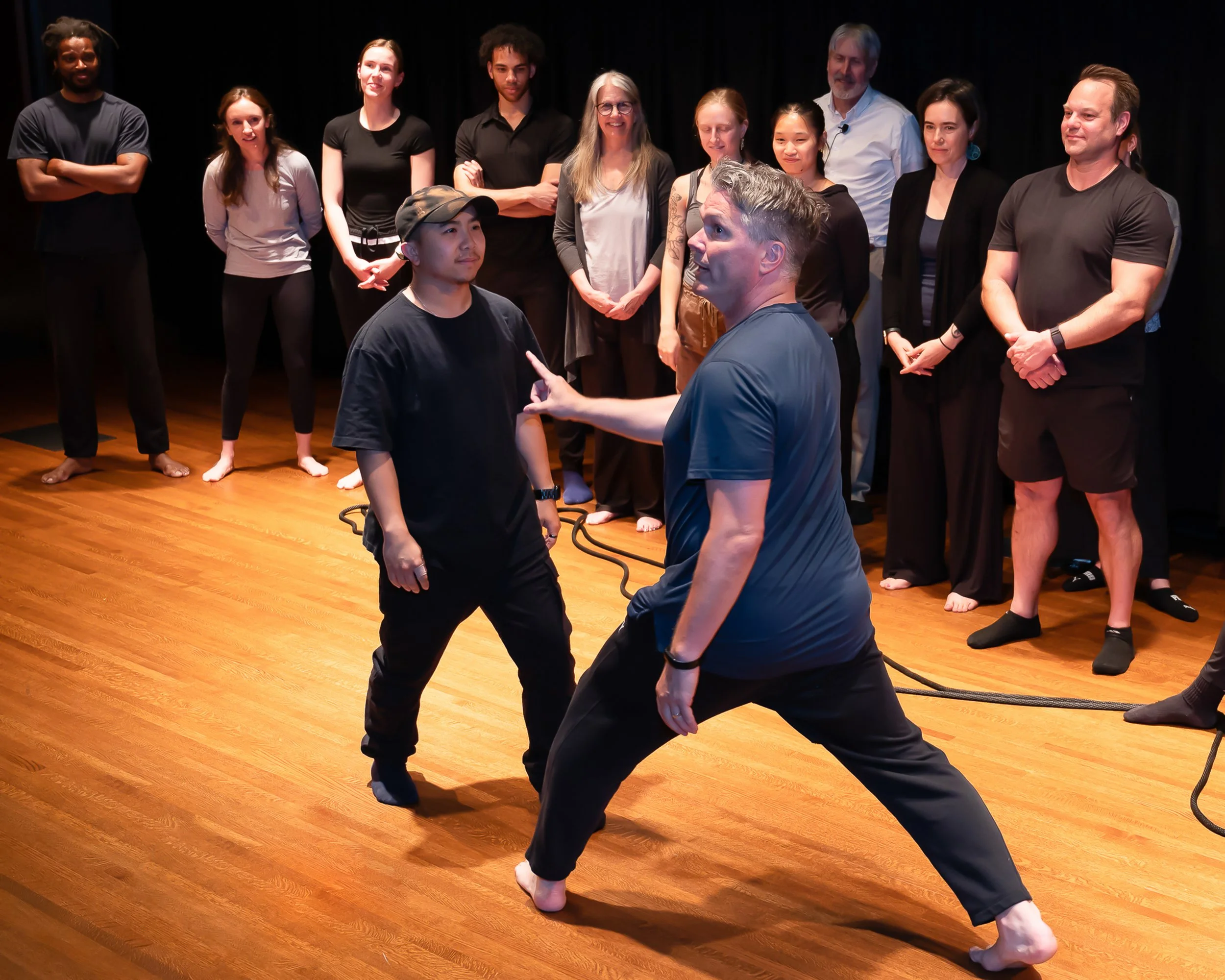 A group of people watching two men performing a martial arts demonstration with ropes on a wooden floor stage.
