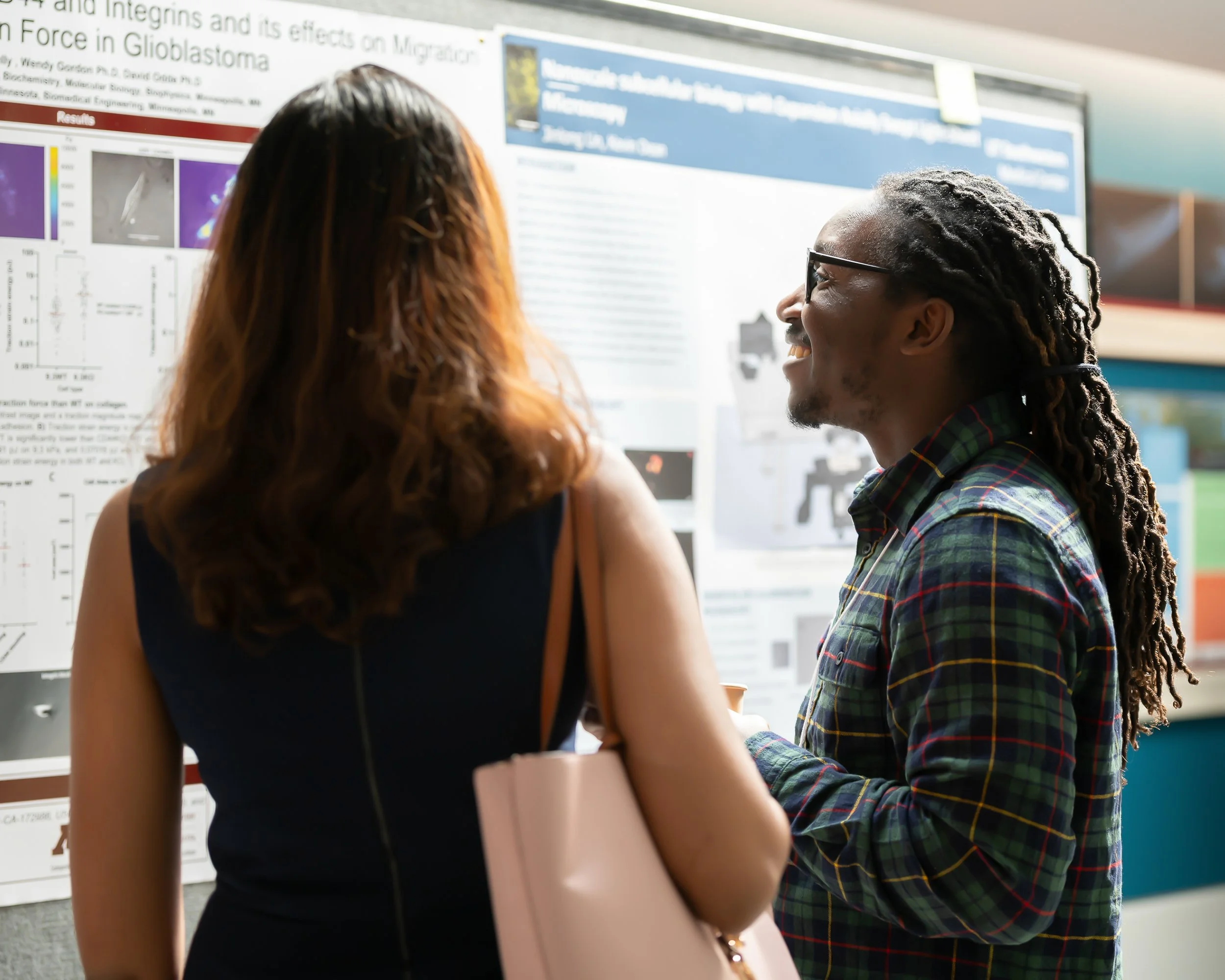 Two people, a woman with long brown hair and a man with long dreadlocks, standing and talking in front of a scientific research poster at a conference or seminar.
