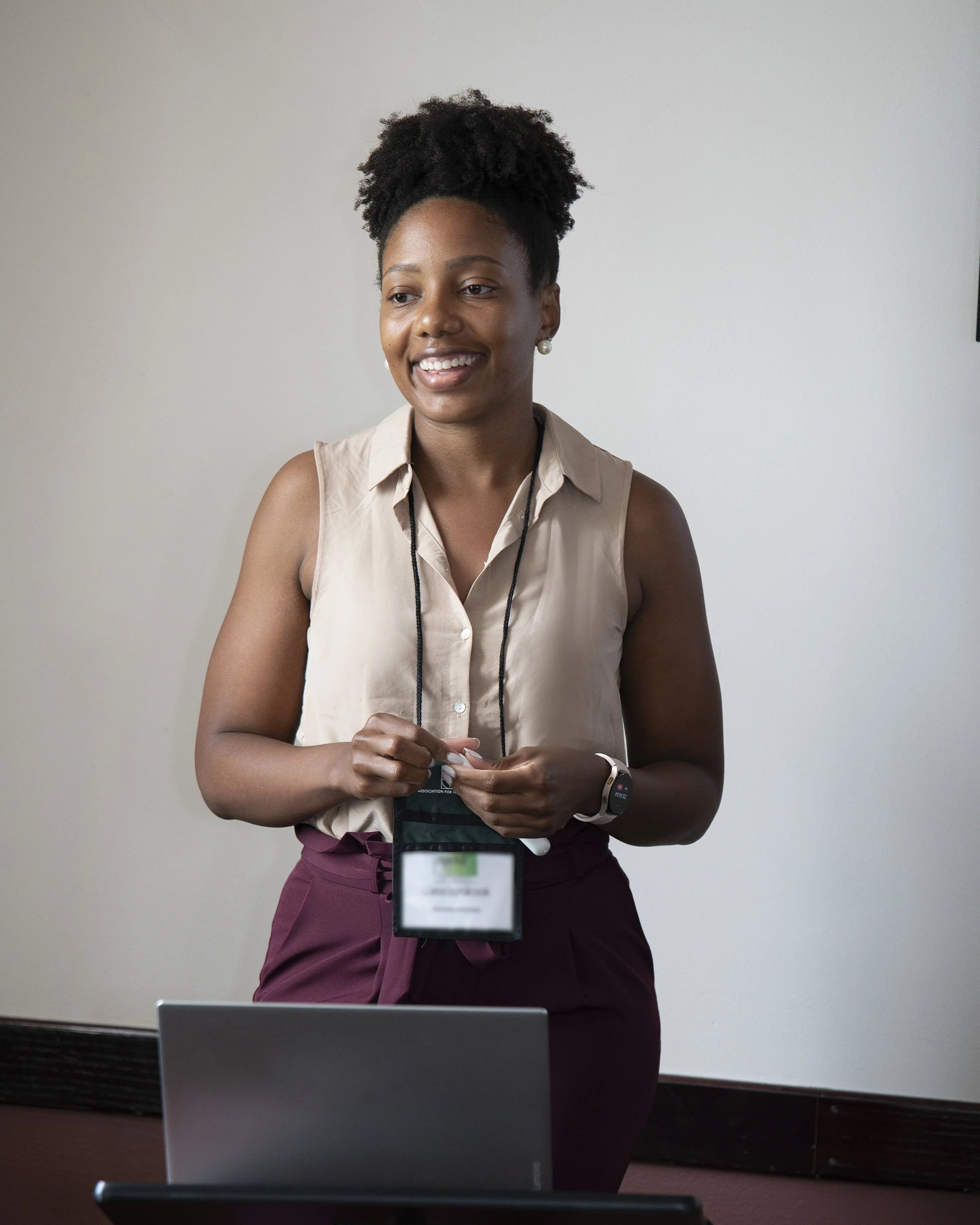 A woman with natural hair smiling, standing in front of a white wall, holding a lanyard with an ID badge.