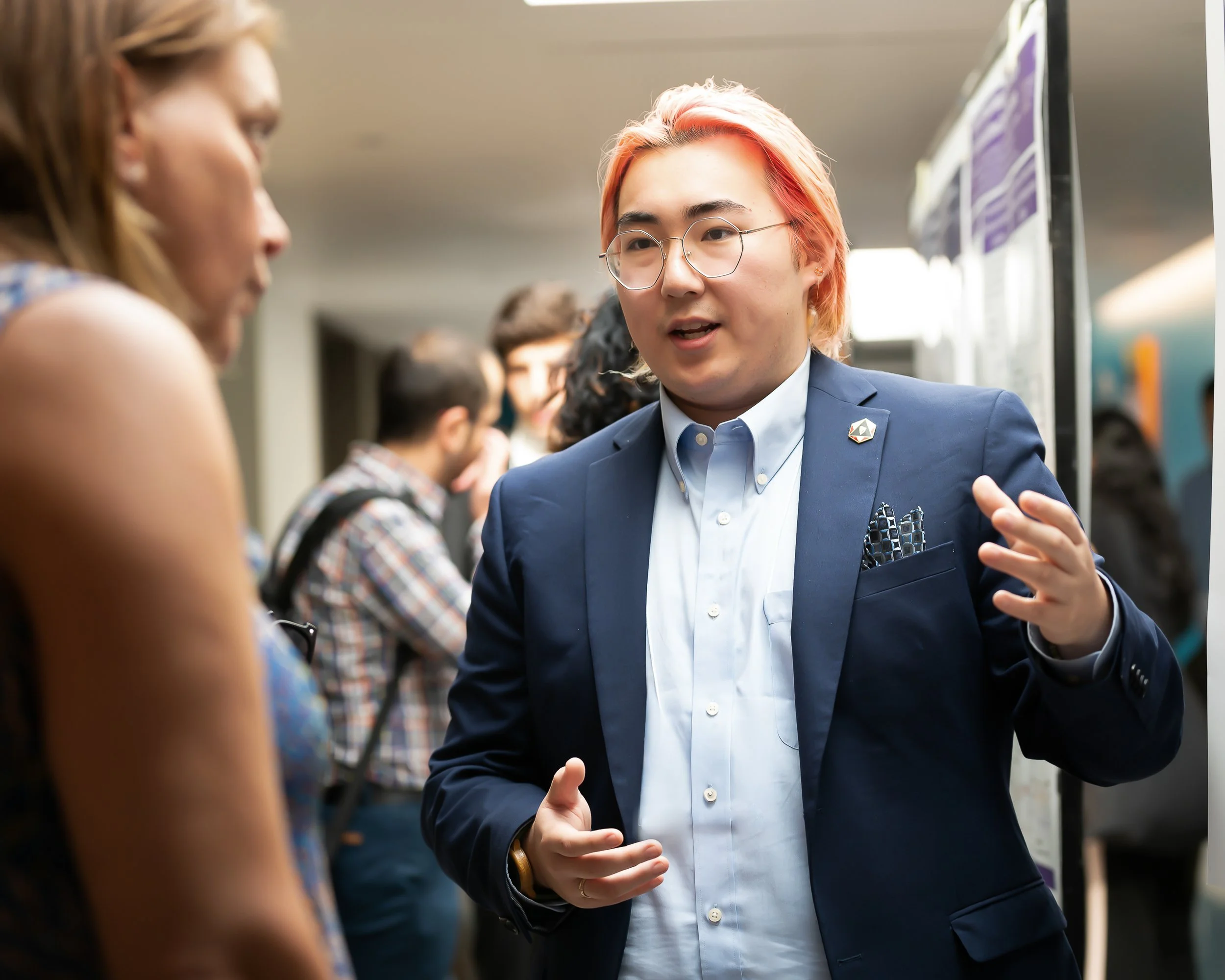 A young man with orange hair and glasses wearing a navy blazer is talking to a woman in a sleeveless top at an indoor event, possibly a conference or networking event.