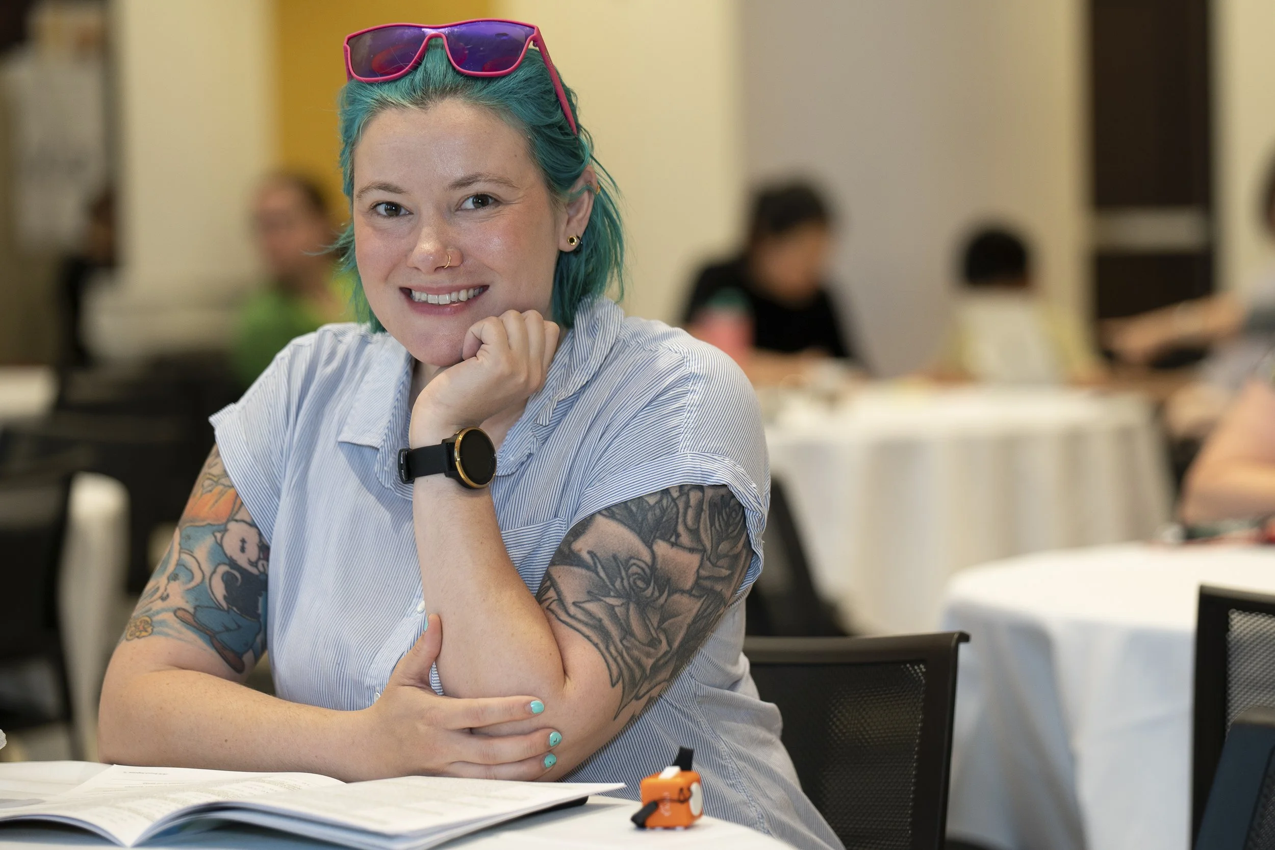 A young woman with short teal hair, wearing pink sunglasses on her head, smiling at the camera. She has tattoos on her arms and is sitting at a table outdoors, with an open book and some small objects in front of her.