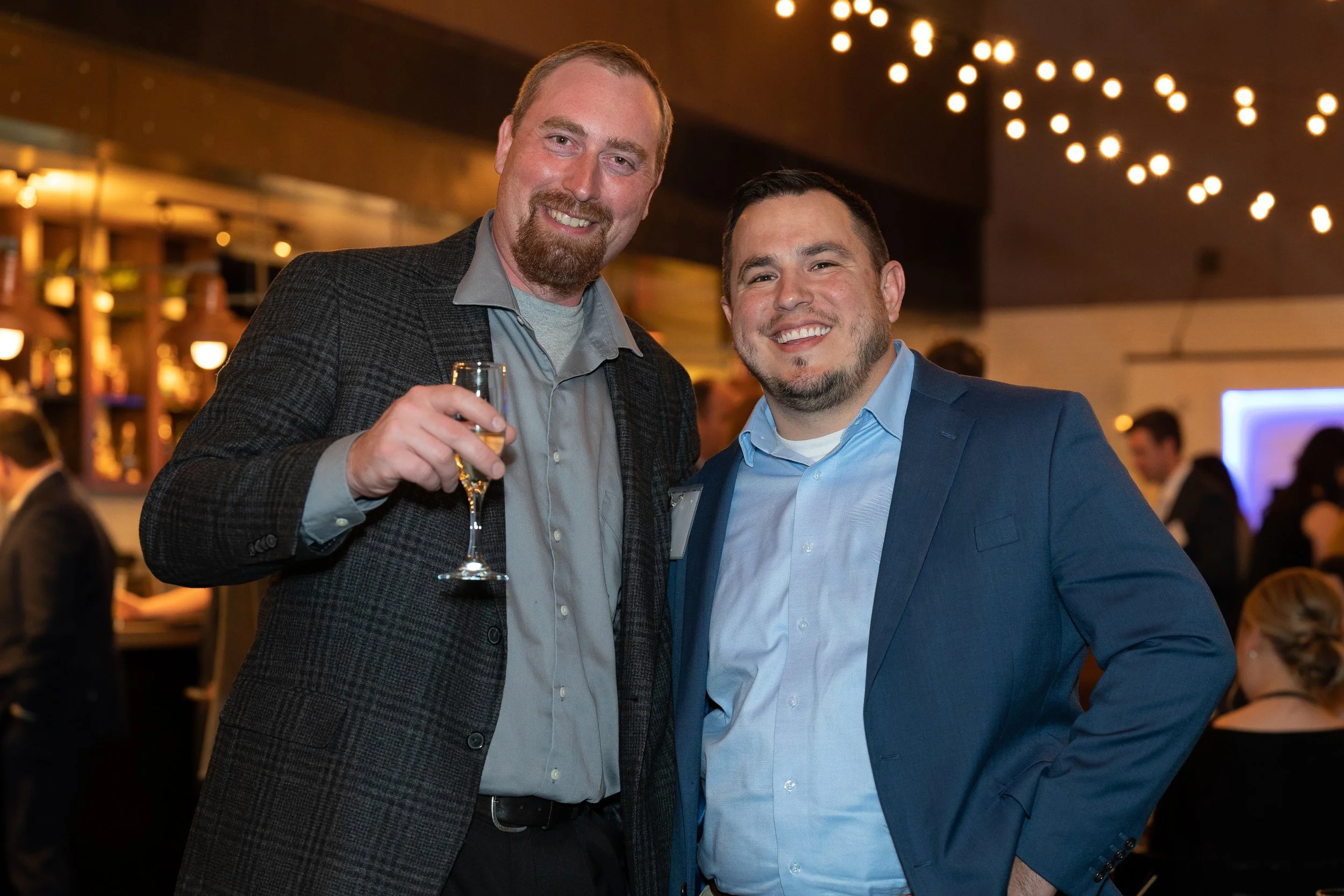 Two men smiling at a social event, one holding a glass of champagne, with warm lighting and a blurred busy background.