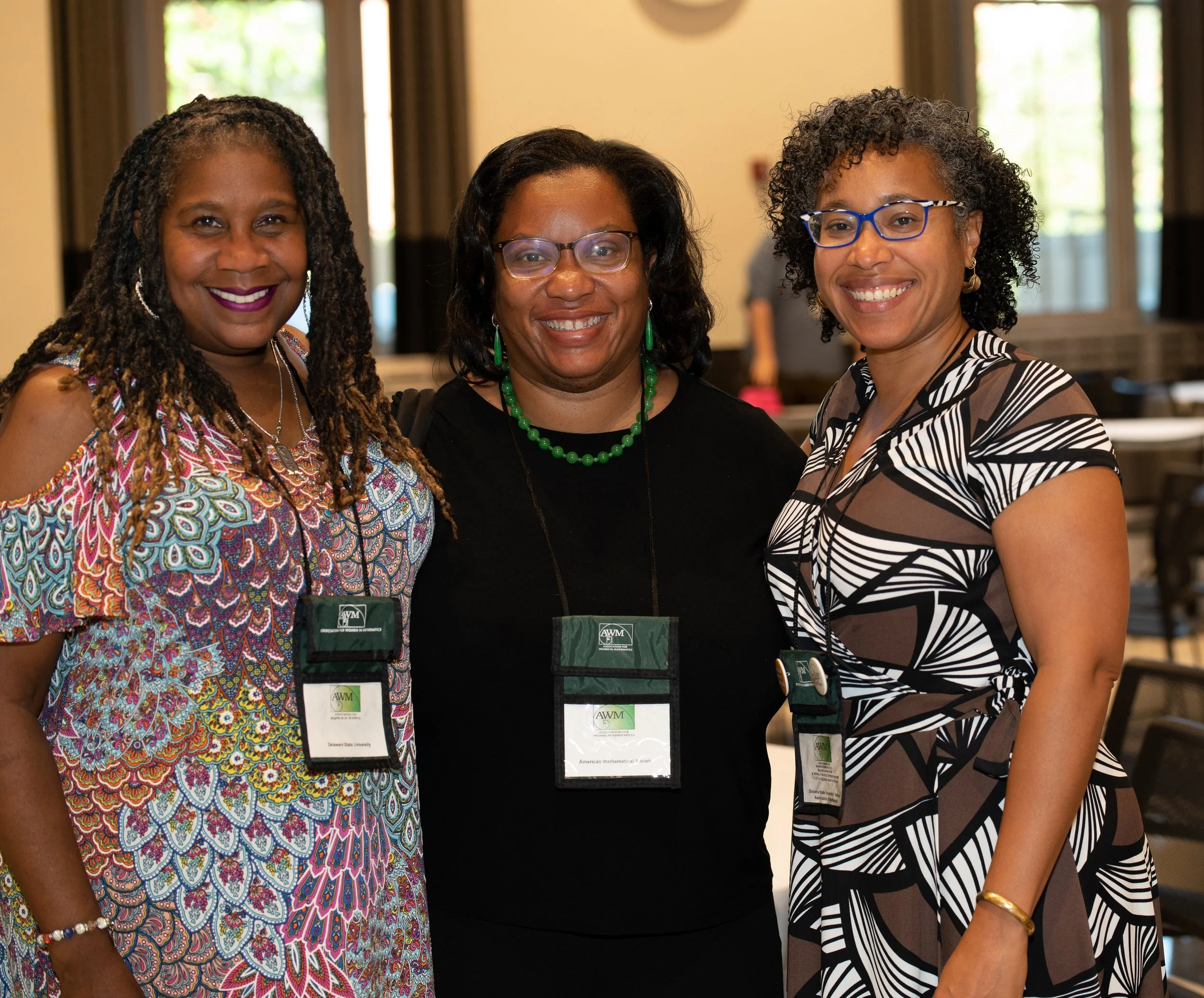 Three women standing close together, smiling, at a conference or event, wearing name tags, with windows and a clock in the background.
