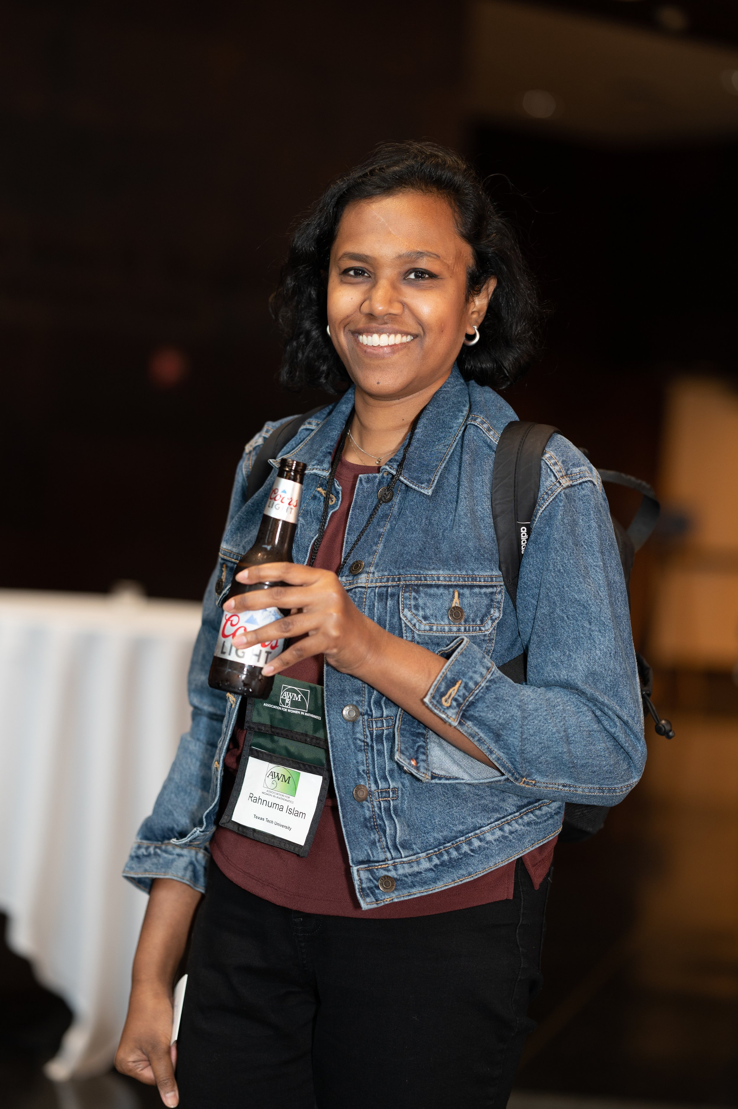 A woman smiling, wearing a denim jacket, holding a beer bottle, and carrying a backpack, standing indoors.