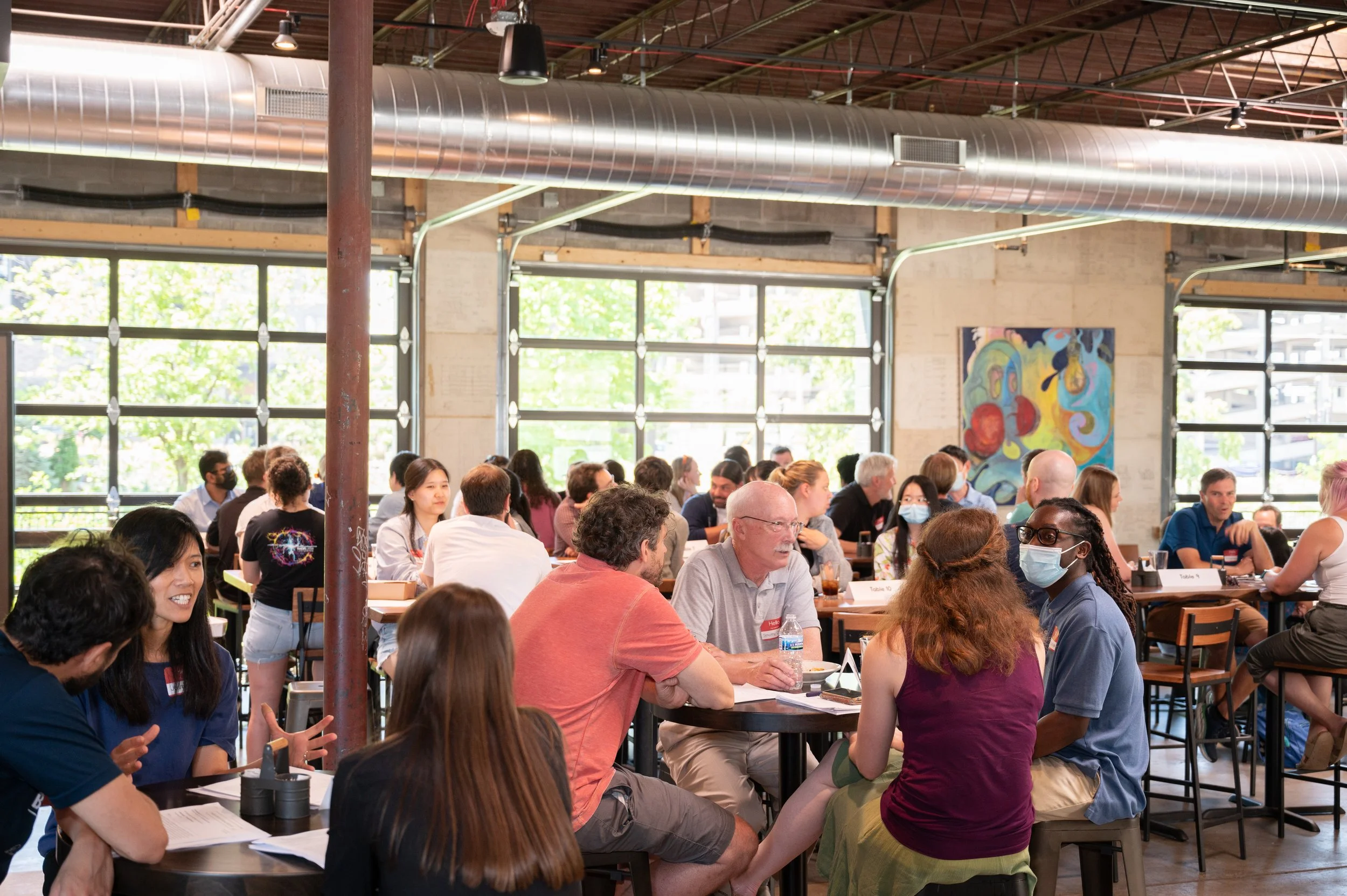People sitting around tables in a bright, industrial-style room with large windows, exposed ductwork, and artwork on the walls.