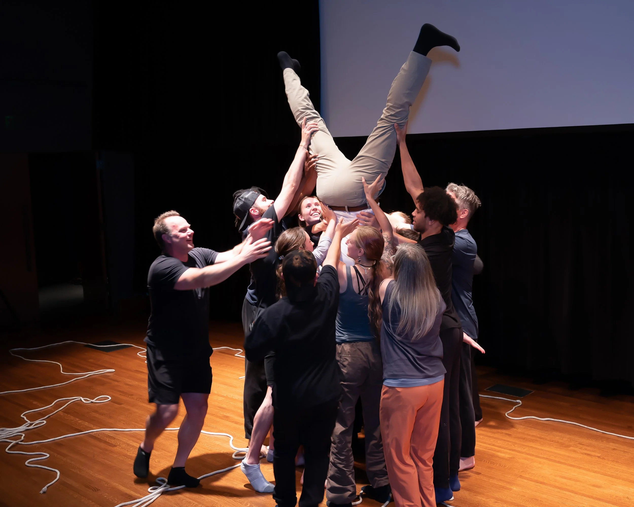 Group of people standing closely together in a circle, lifting a person in beige pants and black shoes in the air during a team activity on a wooden stage with ropes on the floor.