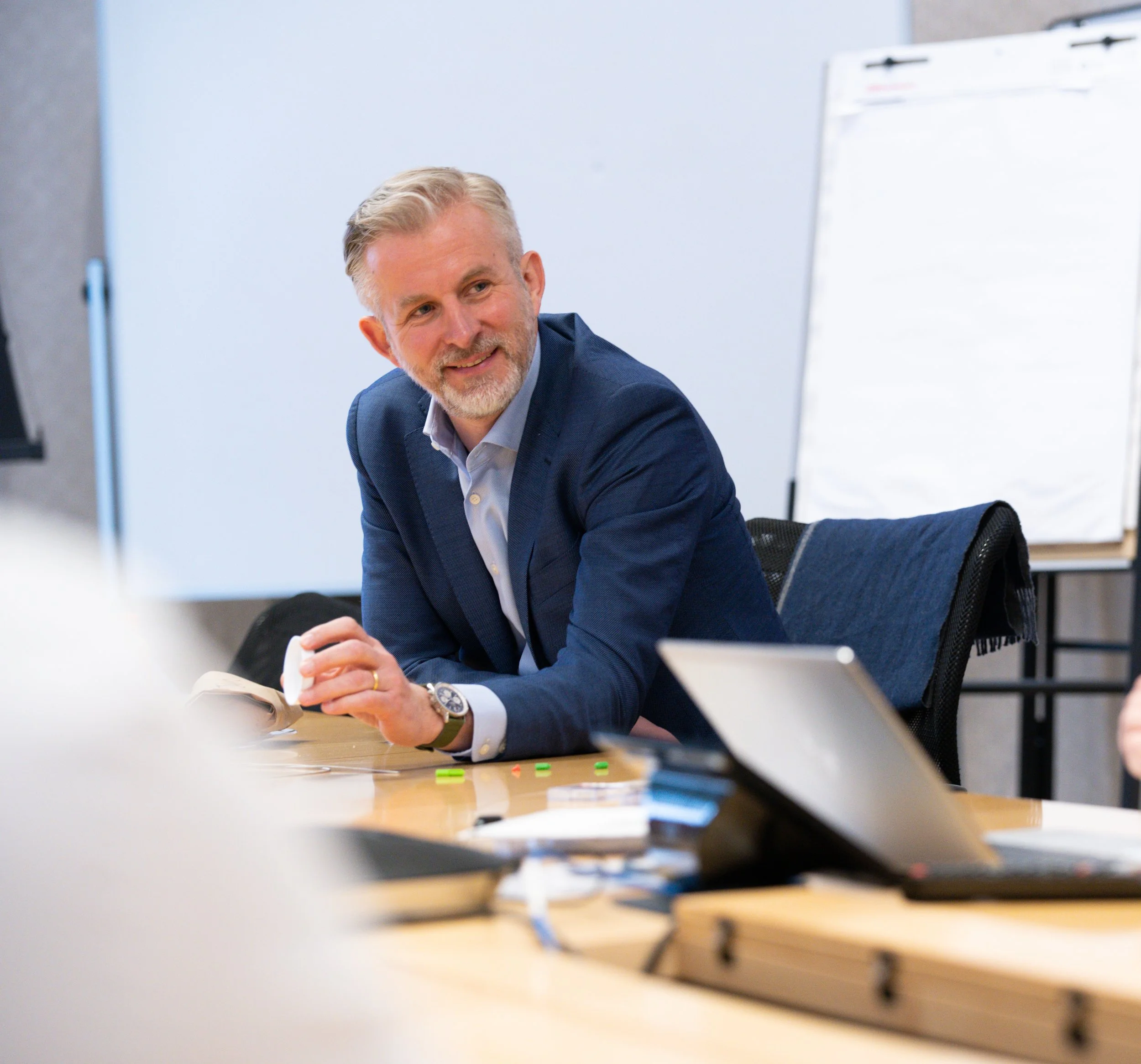 A man with gray hair and a beard, dressed in a blue suit jacket and white shirt, sitting at a desk with his hands clasped, smiling and looking to his left in a modern office setting.