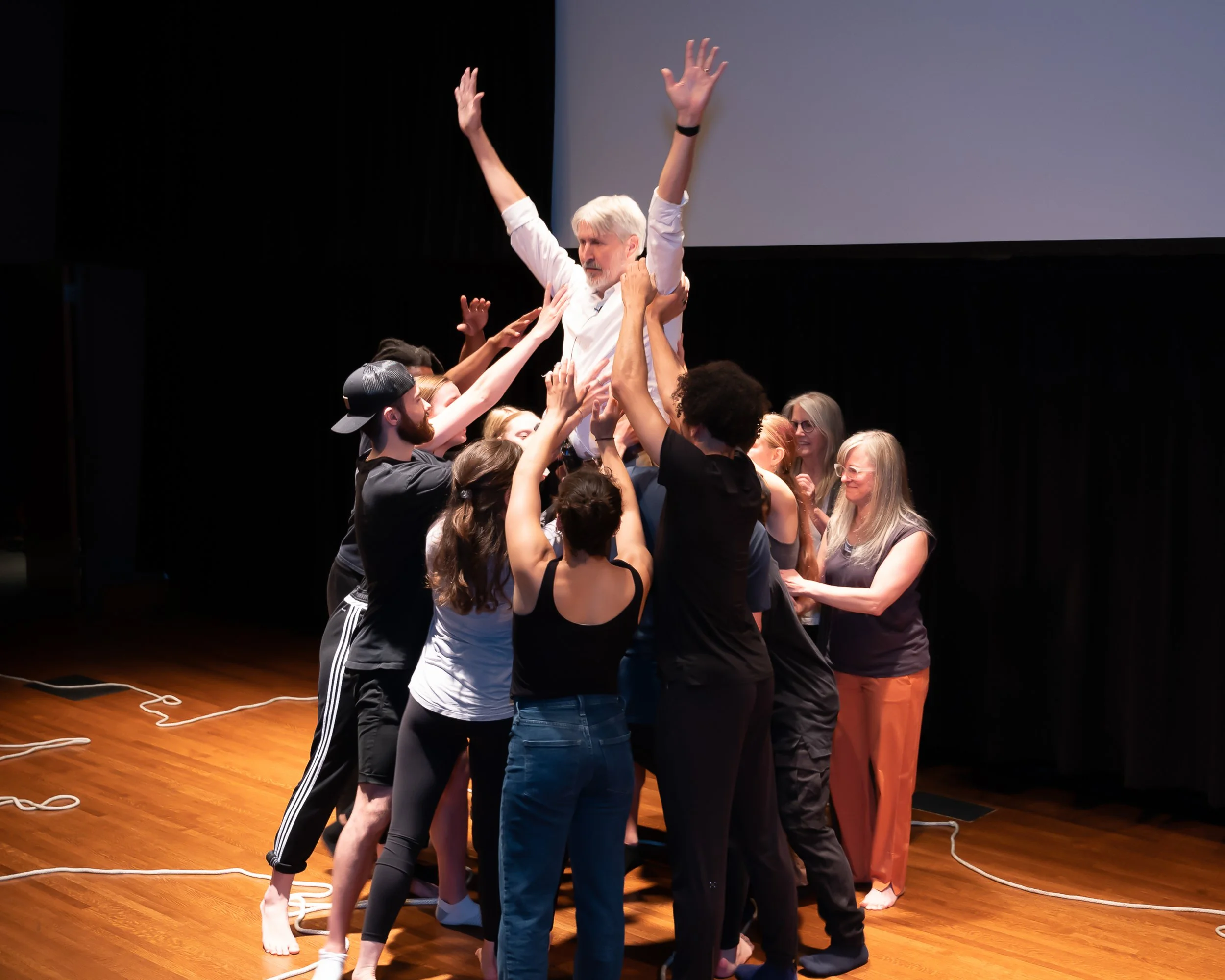 A group of people are participating in a team-building activity on a stage, lifting a man with white hair and beard above their heads. The man has his arms raised. The participants appear engaged and are supporting him from different angles. The stag