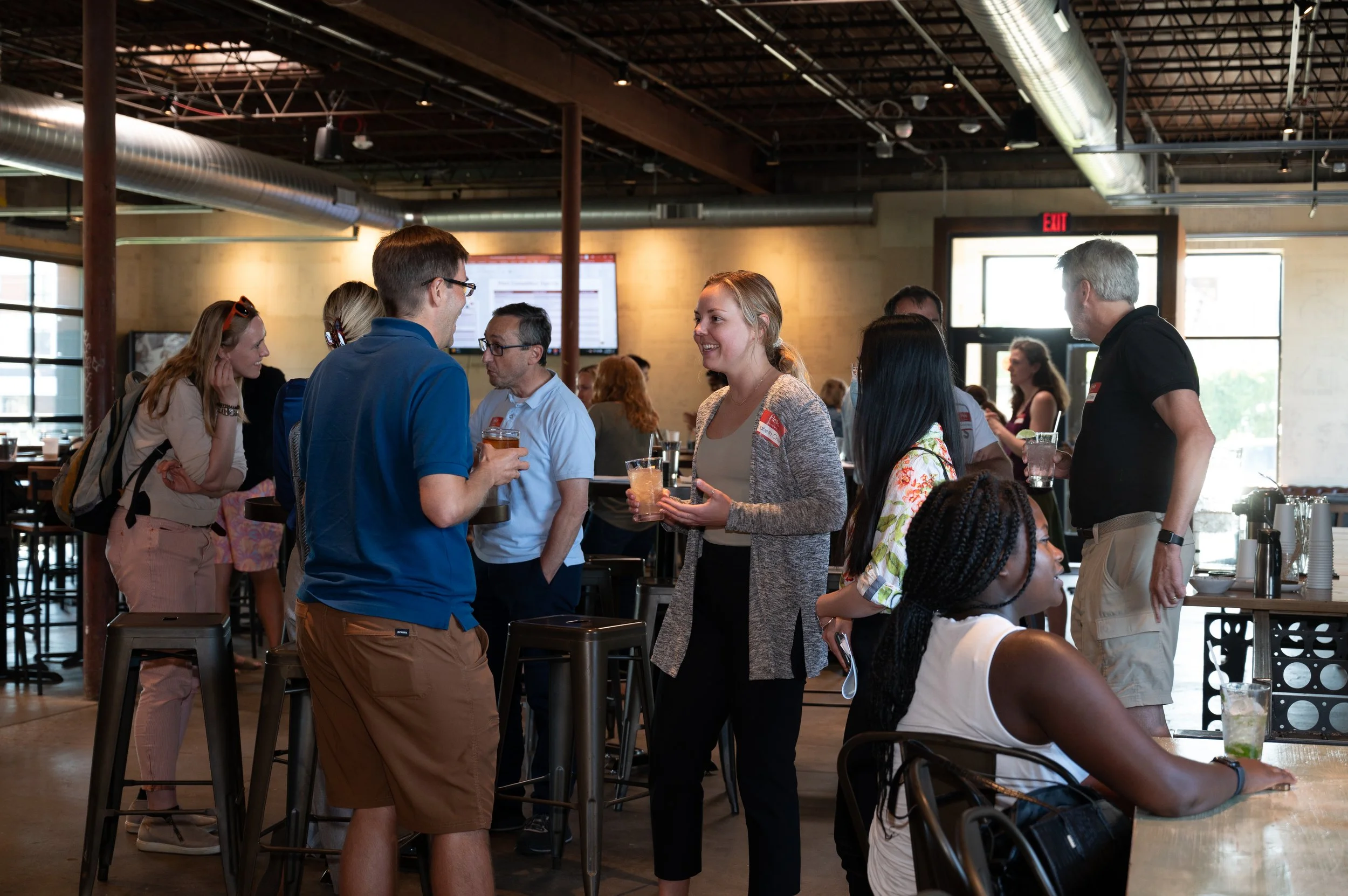Group of people socializing at a bar or cafe, with some holding drinks, inside a modern industrial-style venue with exposed ductwork and large windows.