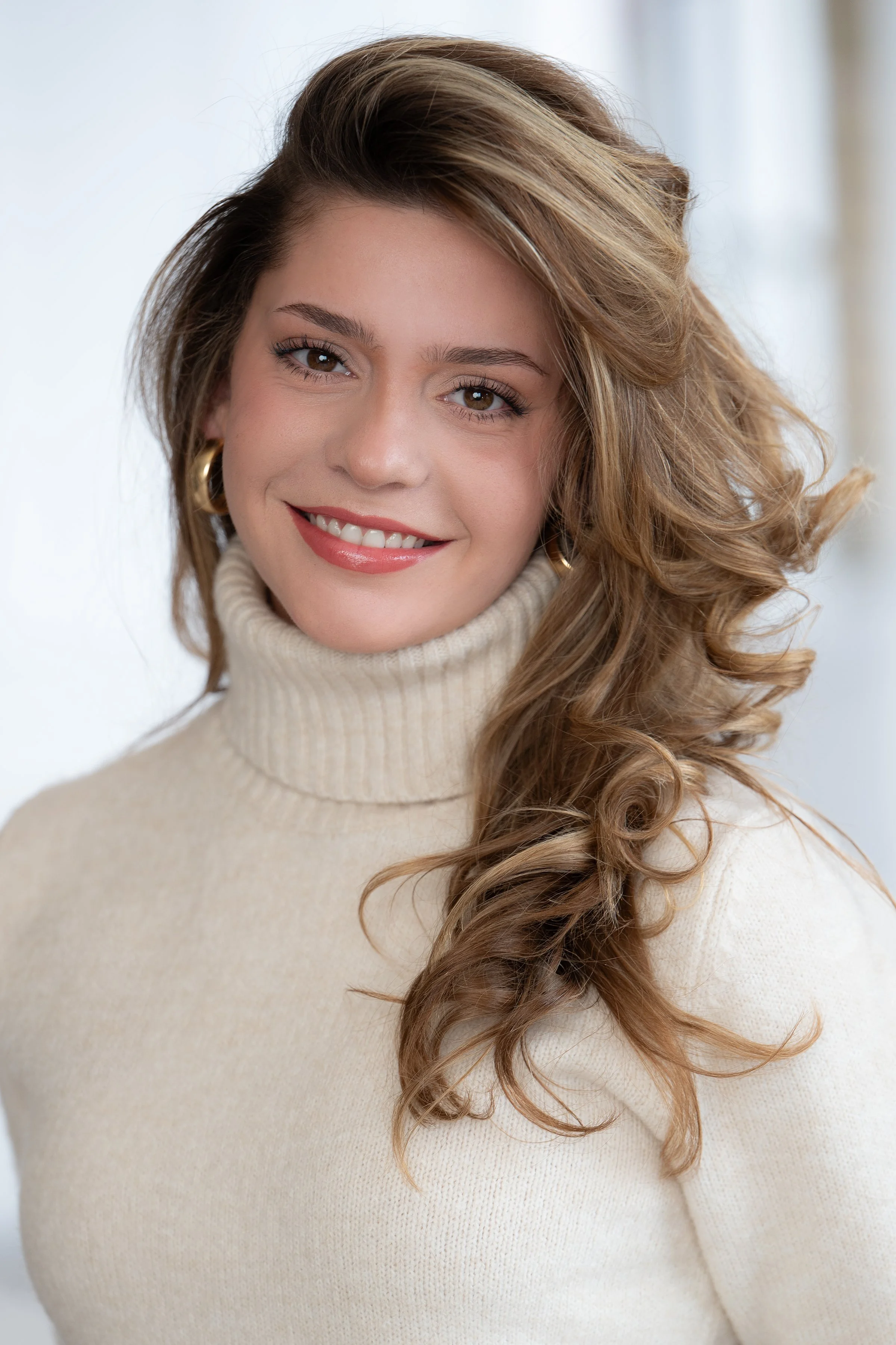 A woman with long, wavy, brown hair, wearing a white turtleneck sweater, smiling with brown eyes and gold hoop earrings, against a plain light background.