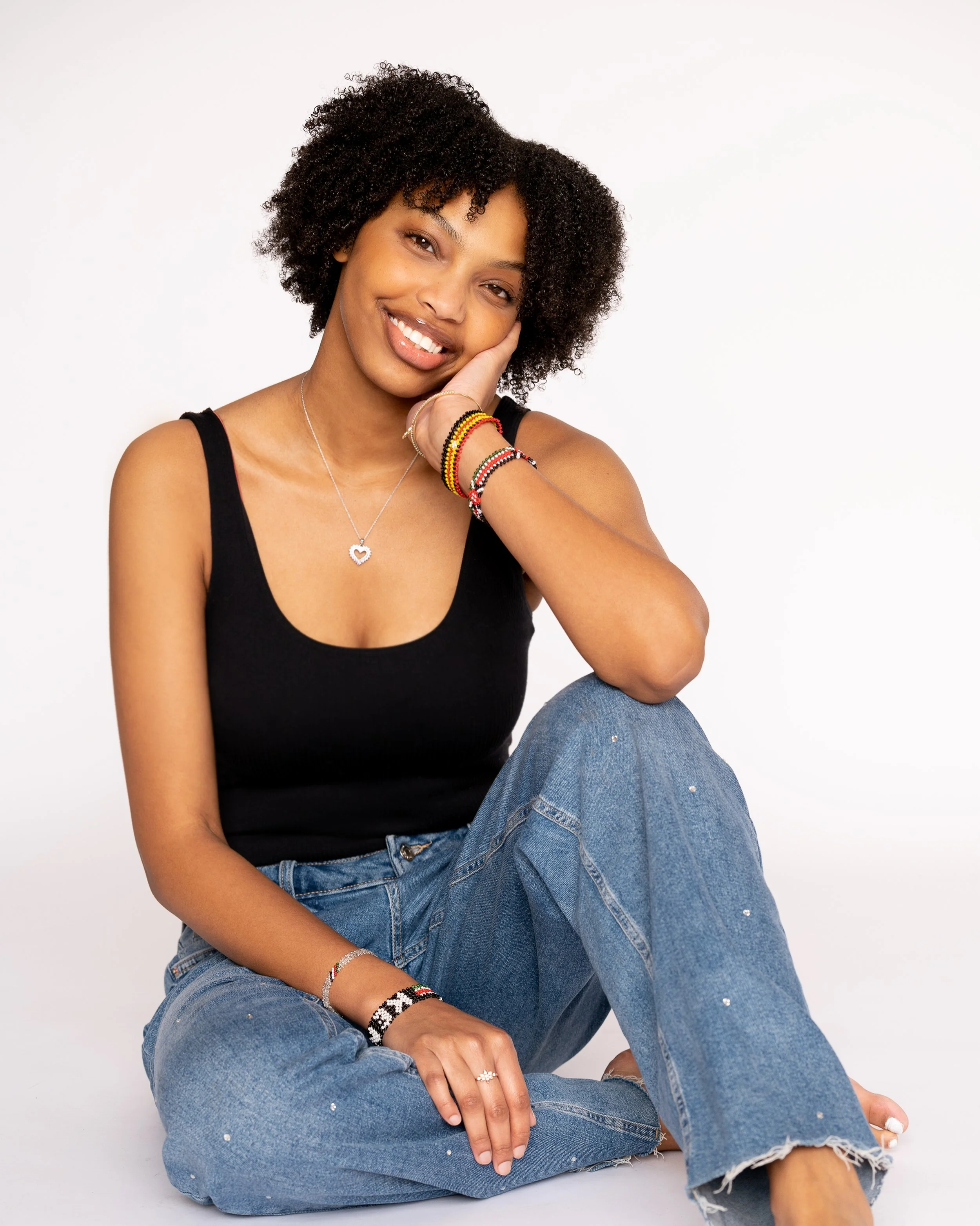 Young woman with curly hair, wearing a black tank top and blue jeans, sitting against a plain white background, smiling and resting her head on her hand with colorful bracelets and a silver necklace.