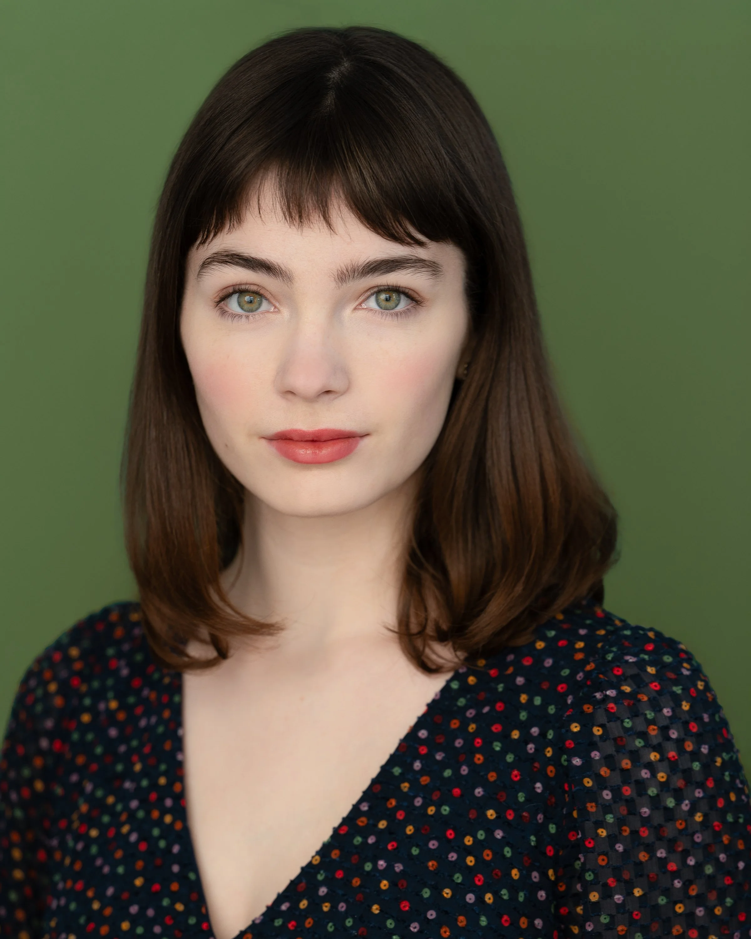 Close-up of a young woman with long brown hair, fair skin, green eyes, wearing a dark top with colorful polka dots, against a green background.