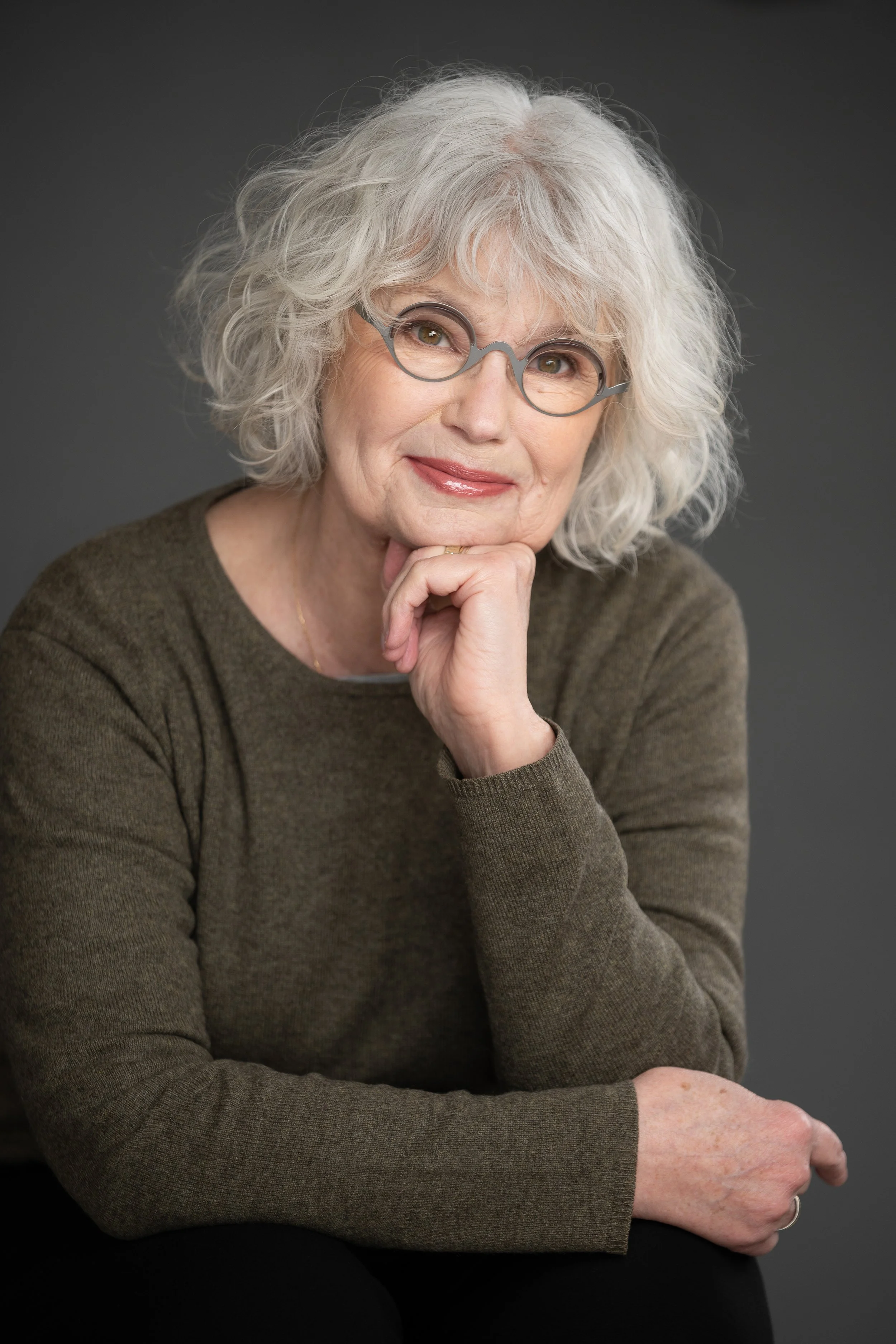 A senior woman with curly gray hair and glasses, wearing a brown sweater, is posing with her hand resting under her chin against a dark gray background.