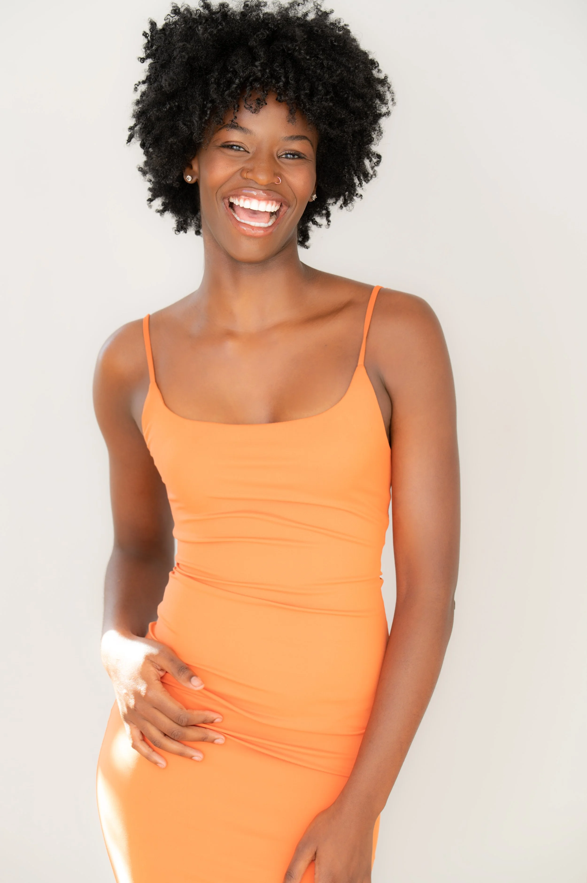 A smiling woman with dark, curly hair wearing an orange dress standing against a plain light-colored background.