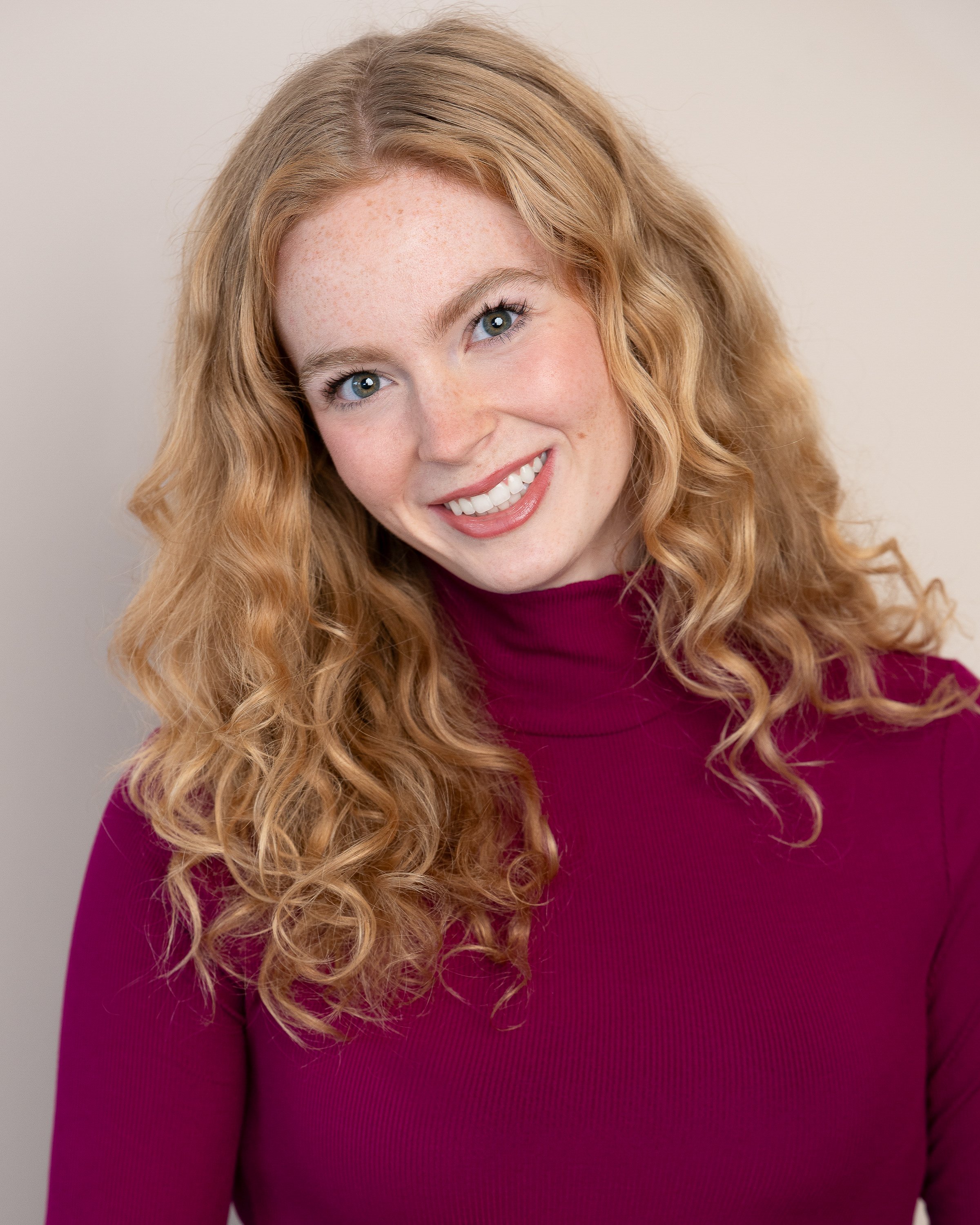 Close-up of a woman with long, curly red hair, wearing a dark pink top, smiling against a plain, light-colored background.