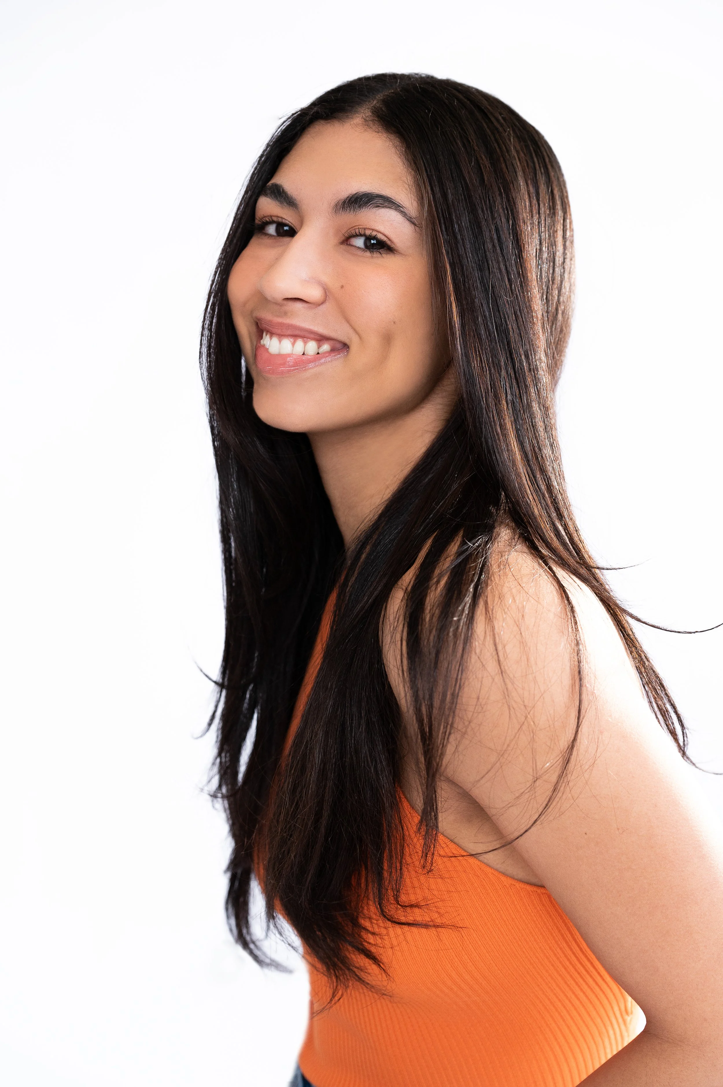 Close-up of a young woman with long dark hair, smiling, wearing an orange sleeveless top against a white background.