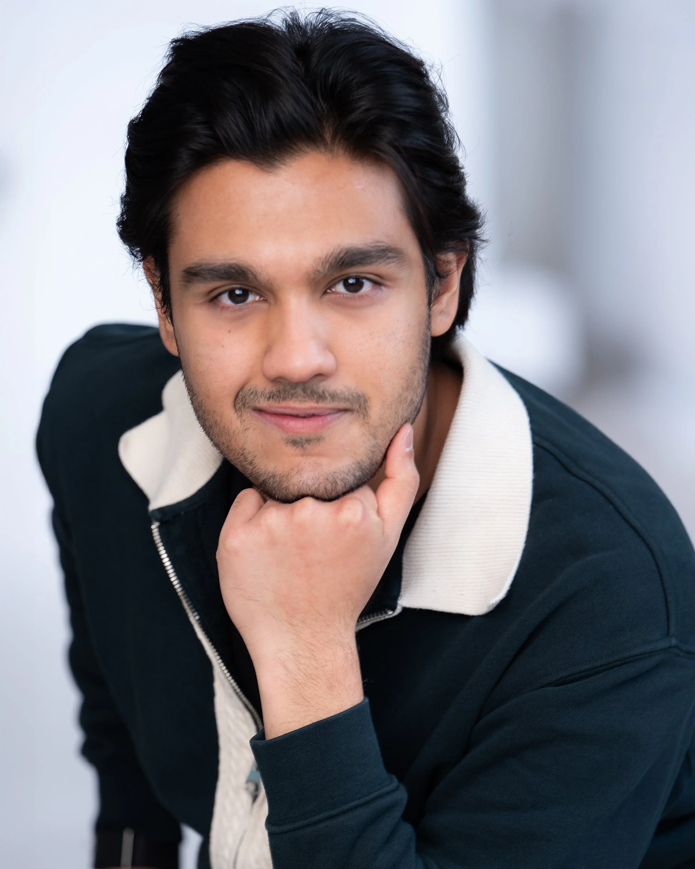 Close-up of a young man with dark hair, resting his chin on his hand, looking directly at the camera with a slight smile, wearing a black and white jacket.