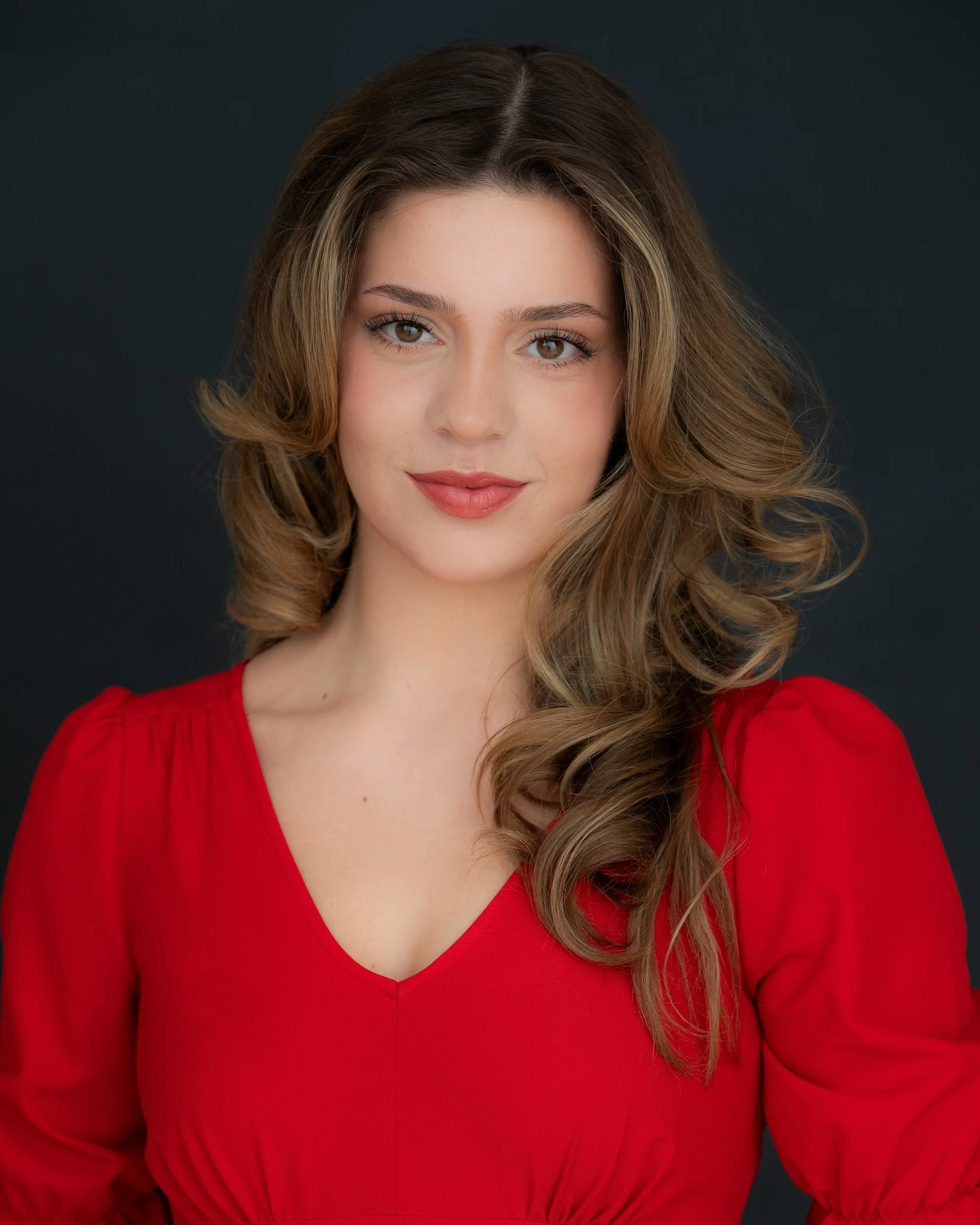 A woman with wavy brown hair, wearing a red top, posing against a dark background.