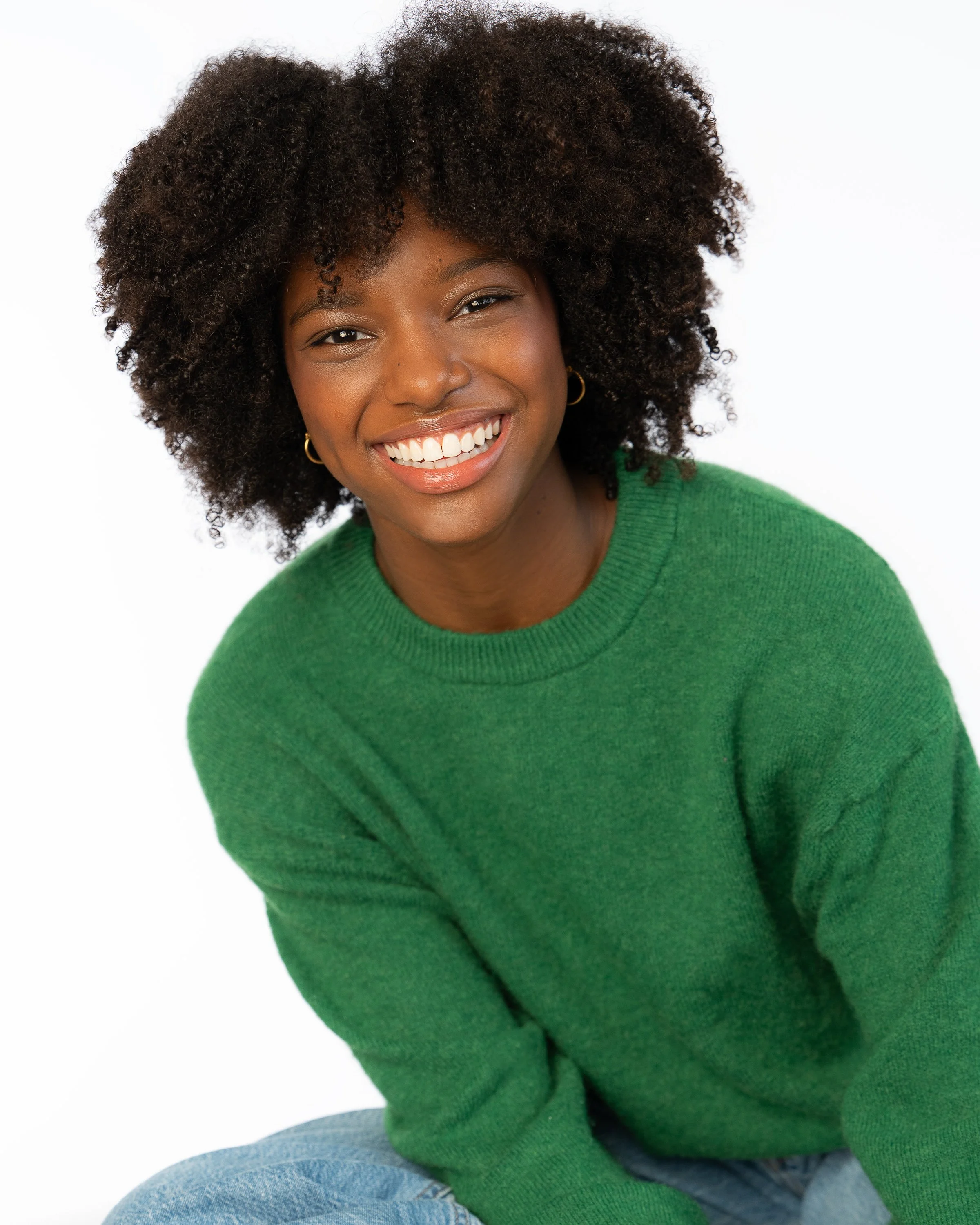 A smiling woman with curly hair wearing a green sweater and blue jeans.