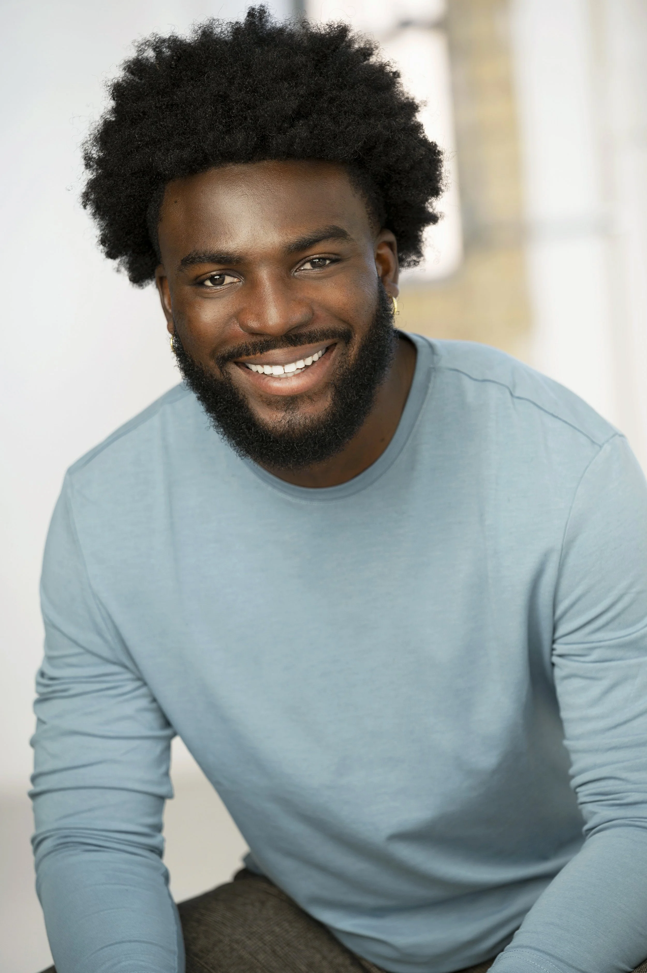 A smiling man with curly black hair, beard, and earrings, wearing a light blue long-sleeve shirt, sitting indoors with a blurred background.