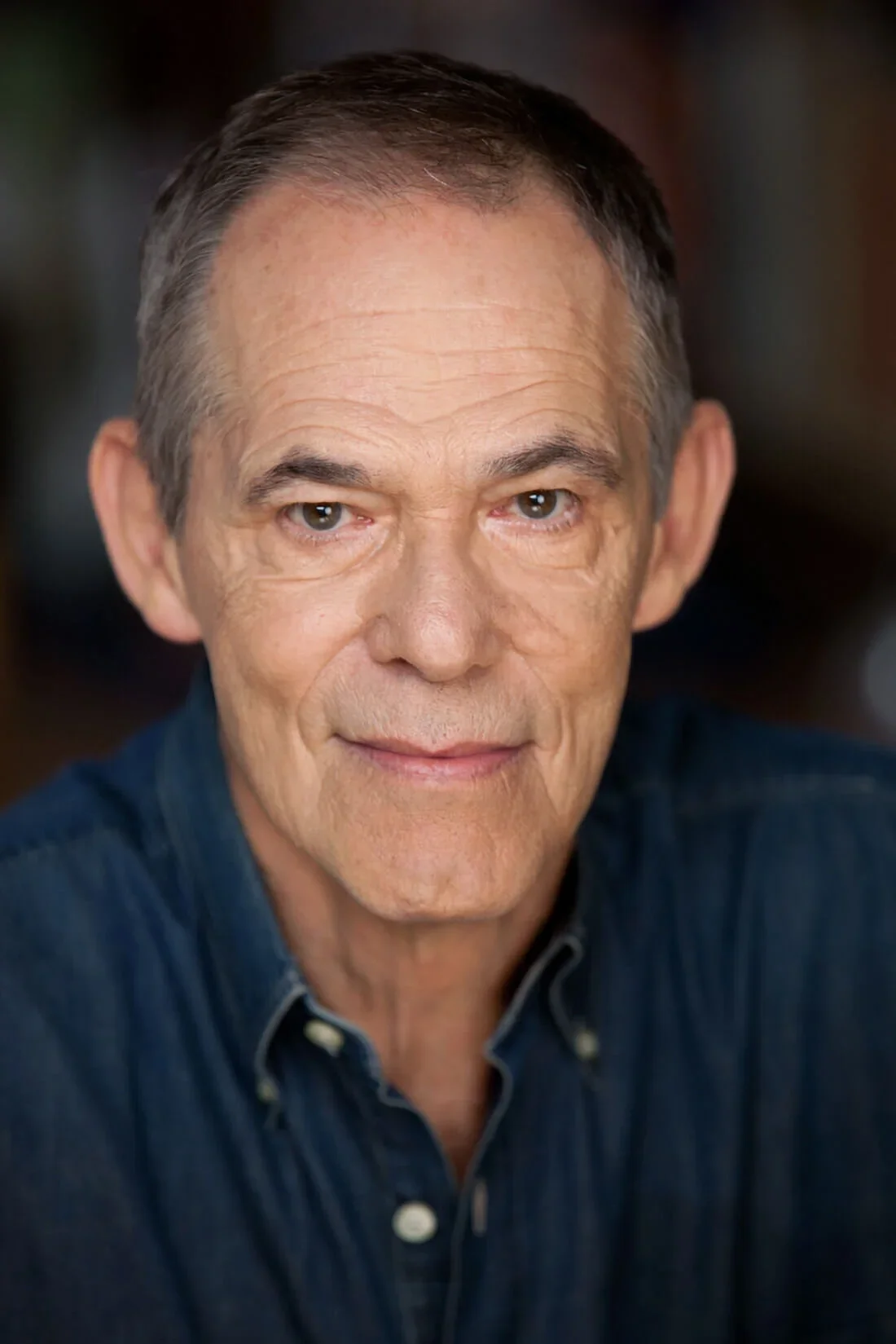 Close-up of an older man with short dark hair, wearing a dark blue button-up shirt, smiling slightly, with a blurred background.