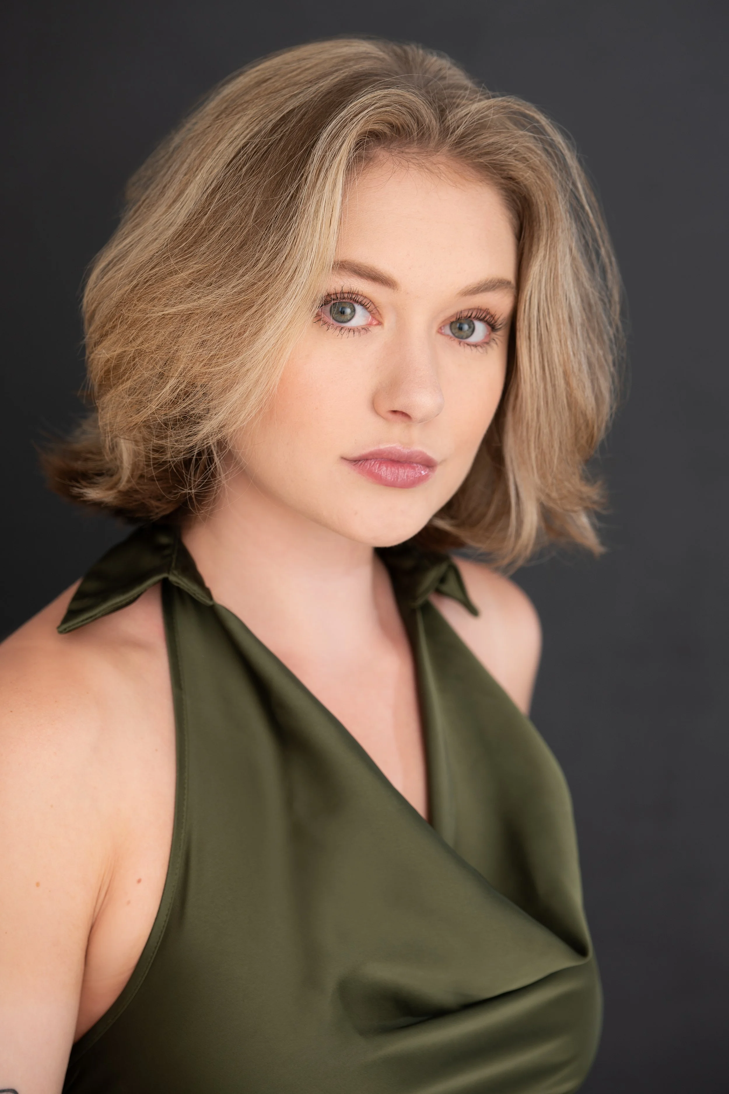 A woman with blonde, shoulder-length wavy hair and blue eyes wearing a dark green satin sleeveless top with a bow on the shoulder, looking at the camera with a neutral expression against a dark gray background.