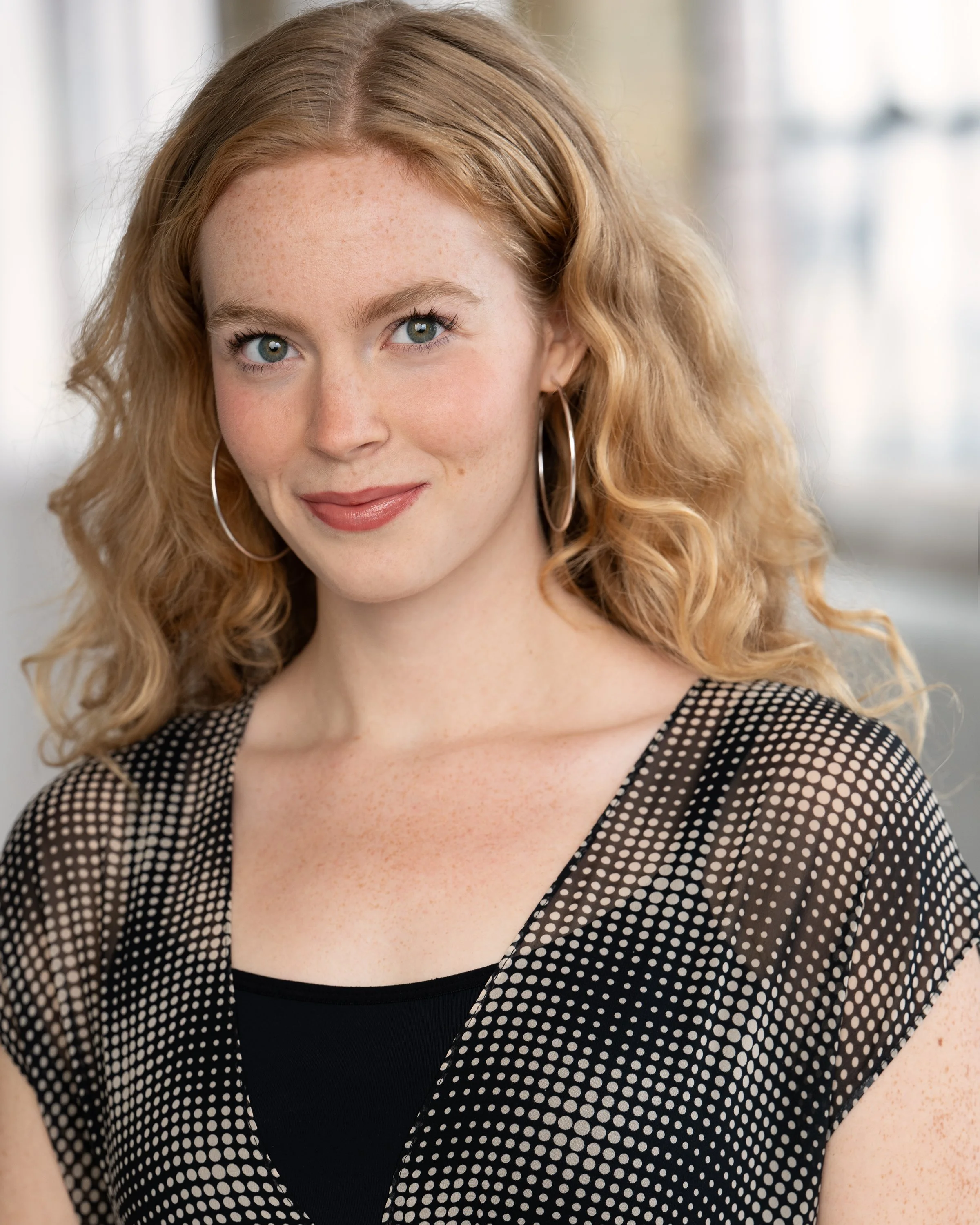 Close-up of a woman with red curly hair, blue eyes, and freckles, wearing hoop earrings and a black and white polka dot top.