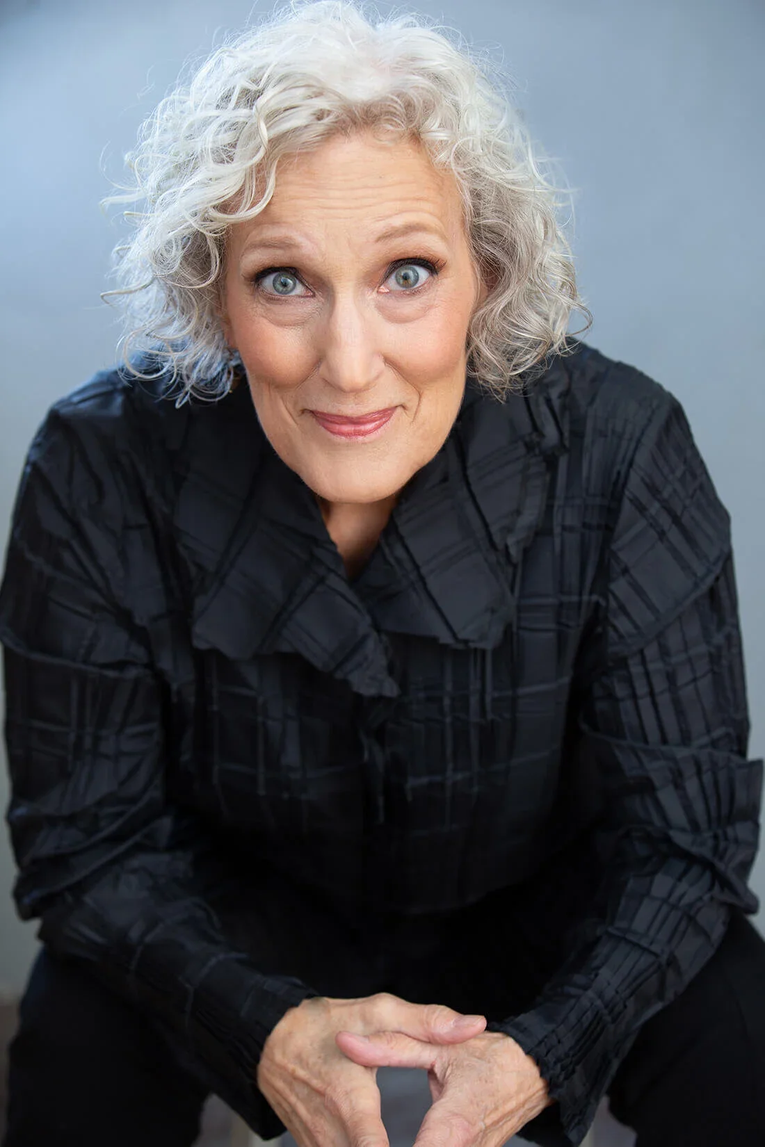 A woman with silver, curly hair and blue eyes wearing a black checked shirt, smiling at the camera against a light grey background.