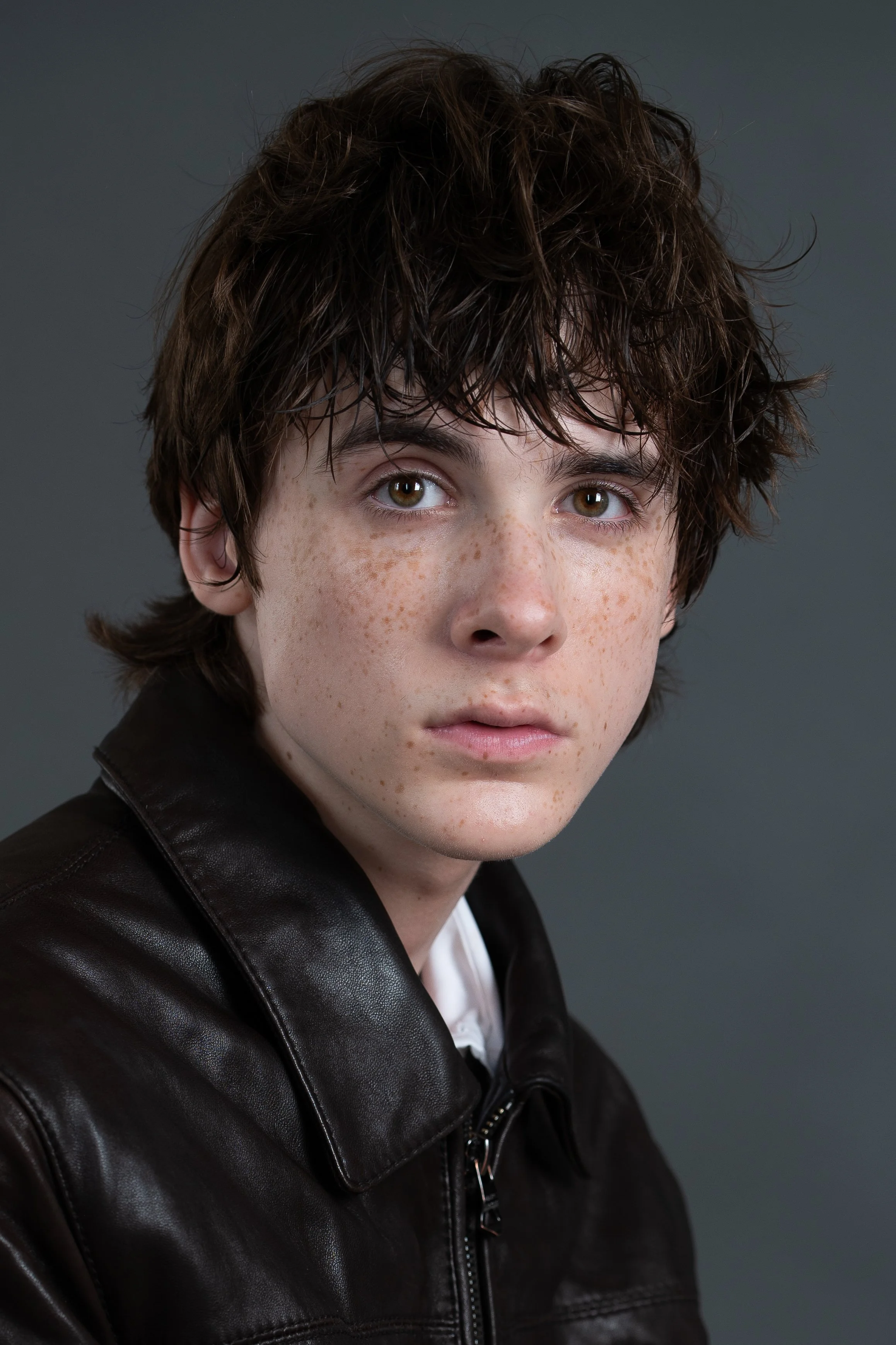 Close-up portrait of a young man with brown, curly hair, freckles, hazel eyes, wearing a black leather jacket against a plain gray background.