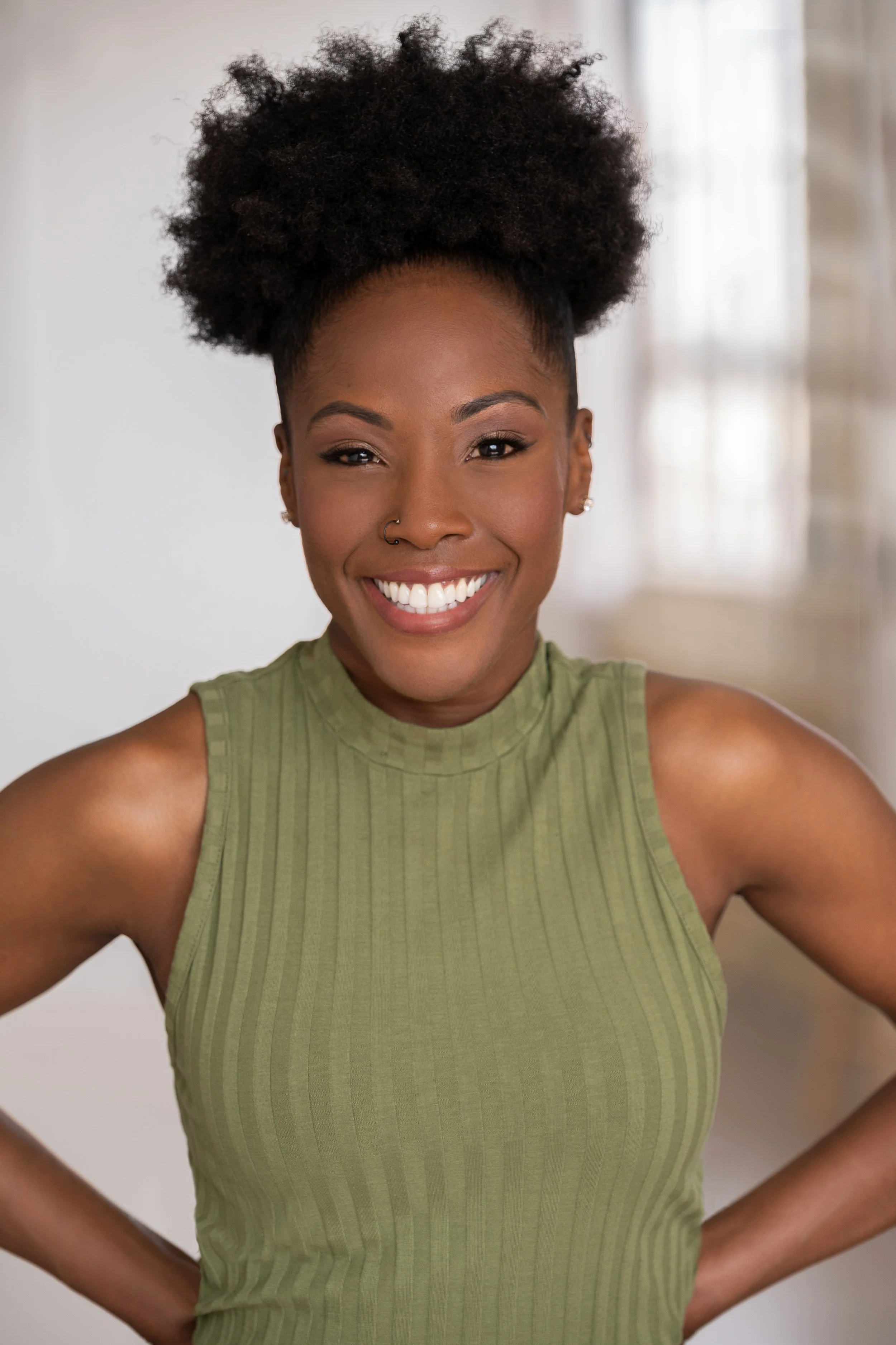 A woman with natural black curly hair styled in an updo, wearing a green sleeveless ribbed top, smiling, standing in front of a blurred, light background.