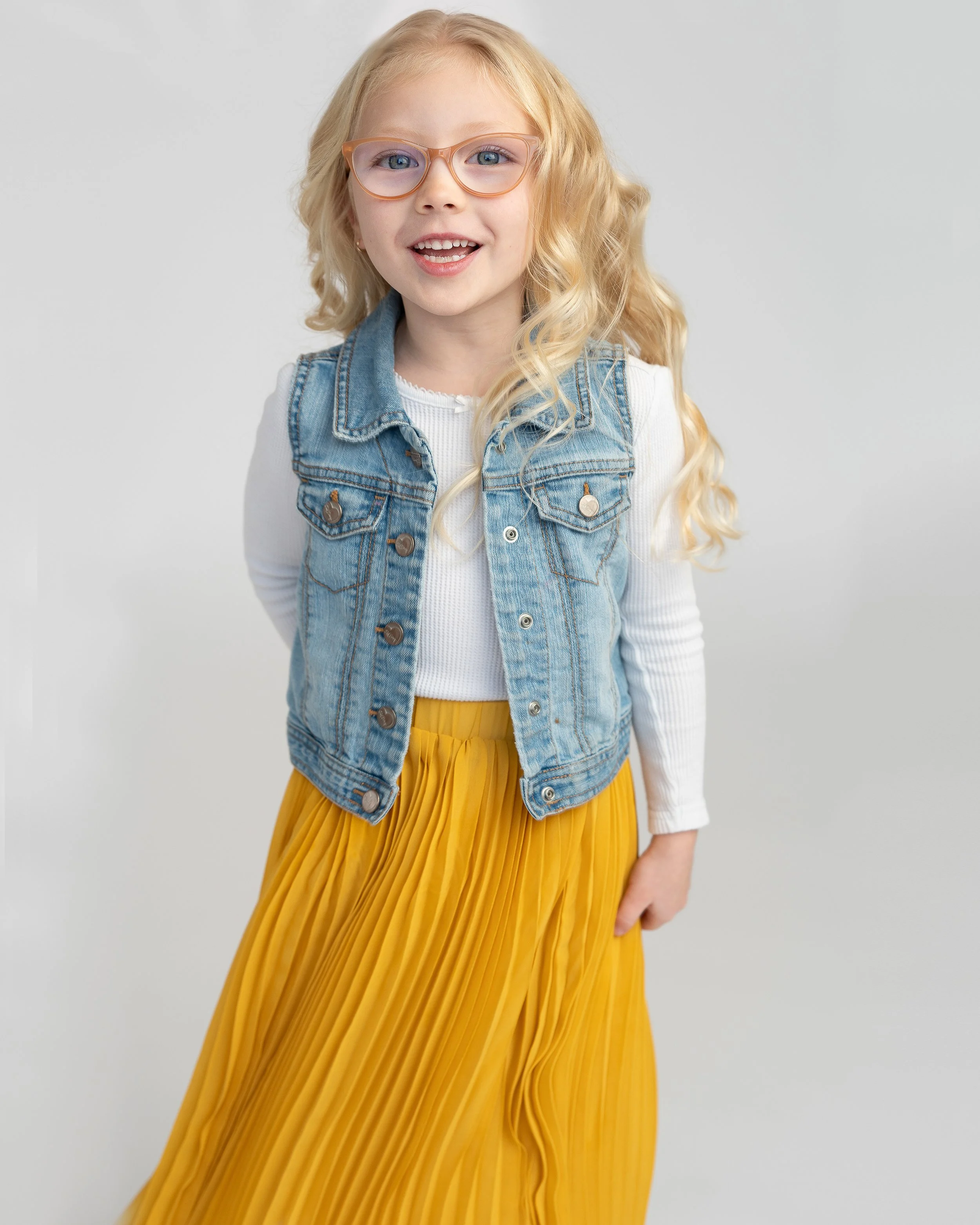 A young girl with blonde hair and blue eyes wearing glasses, a denim vest over a white long-sleeve shirt, and a yellow pleated skirt, smiling against a plain light gray background.
