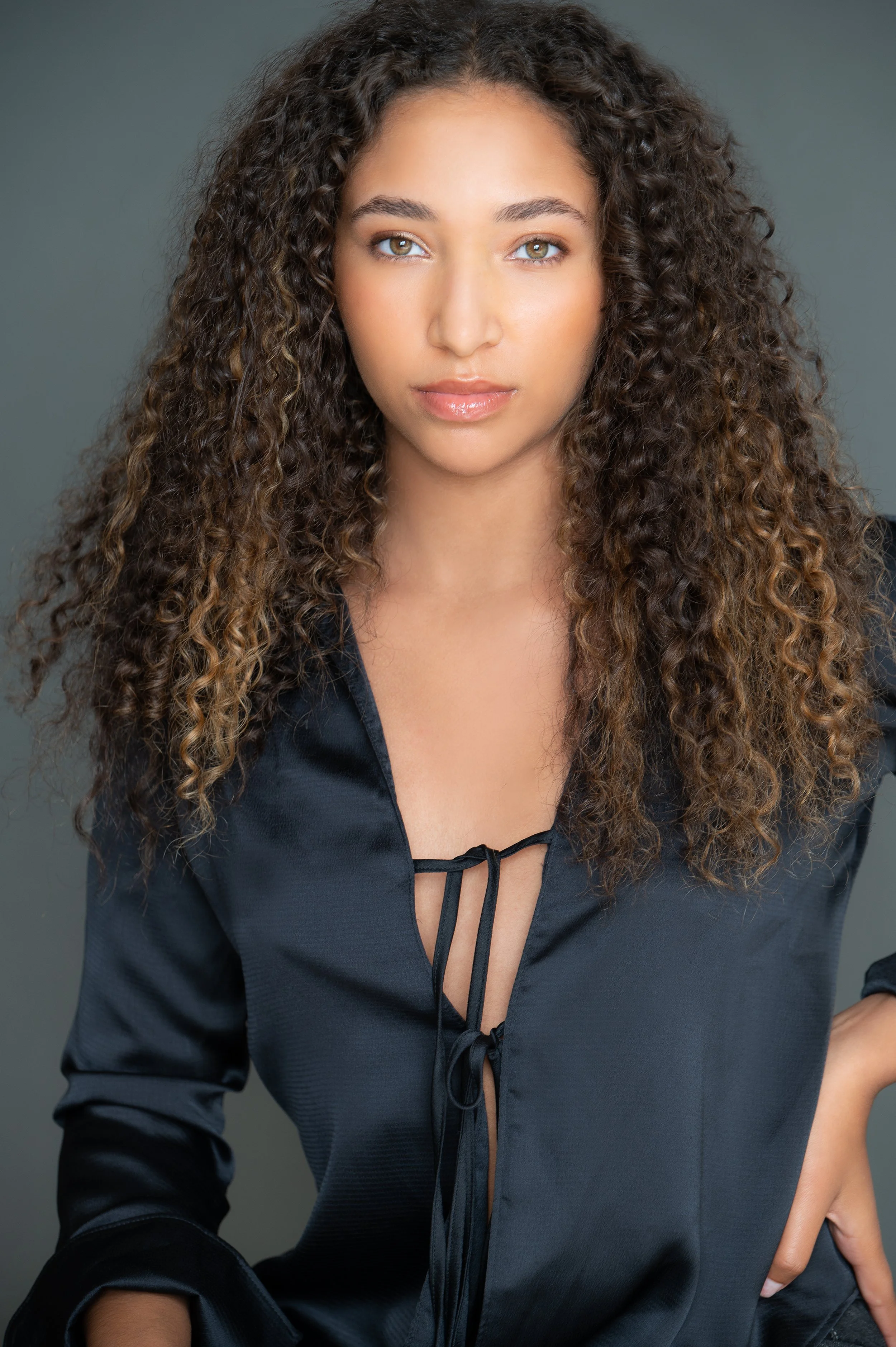 A young woman with long curly hair and light brown eyes wearing a black satin top with a lace-up front against a gray background.