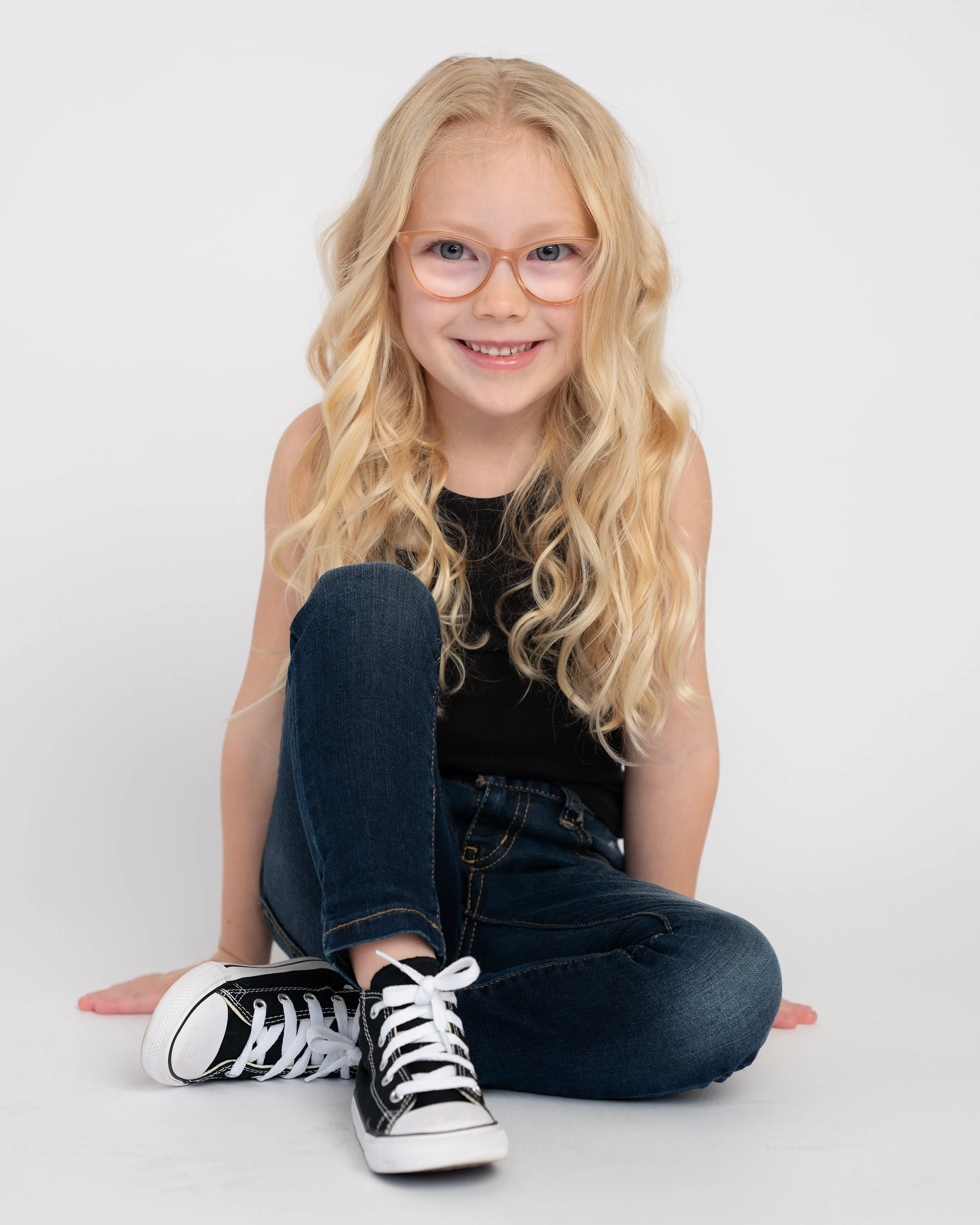 Young girl with long blonde curly hair wearing pink glasses, black tank top, dark jeans, and black sneakers with white laces, sitting on the white floor with a plain white background.