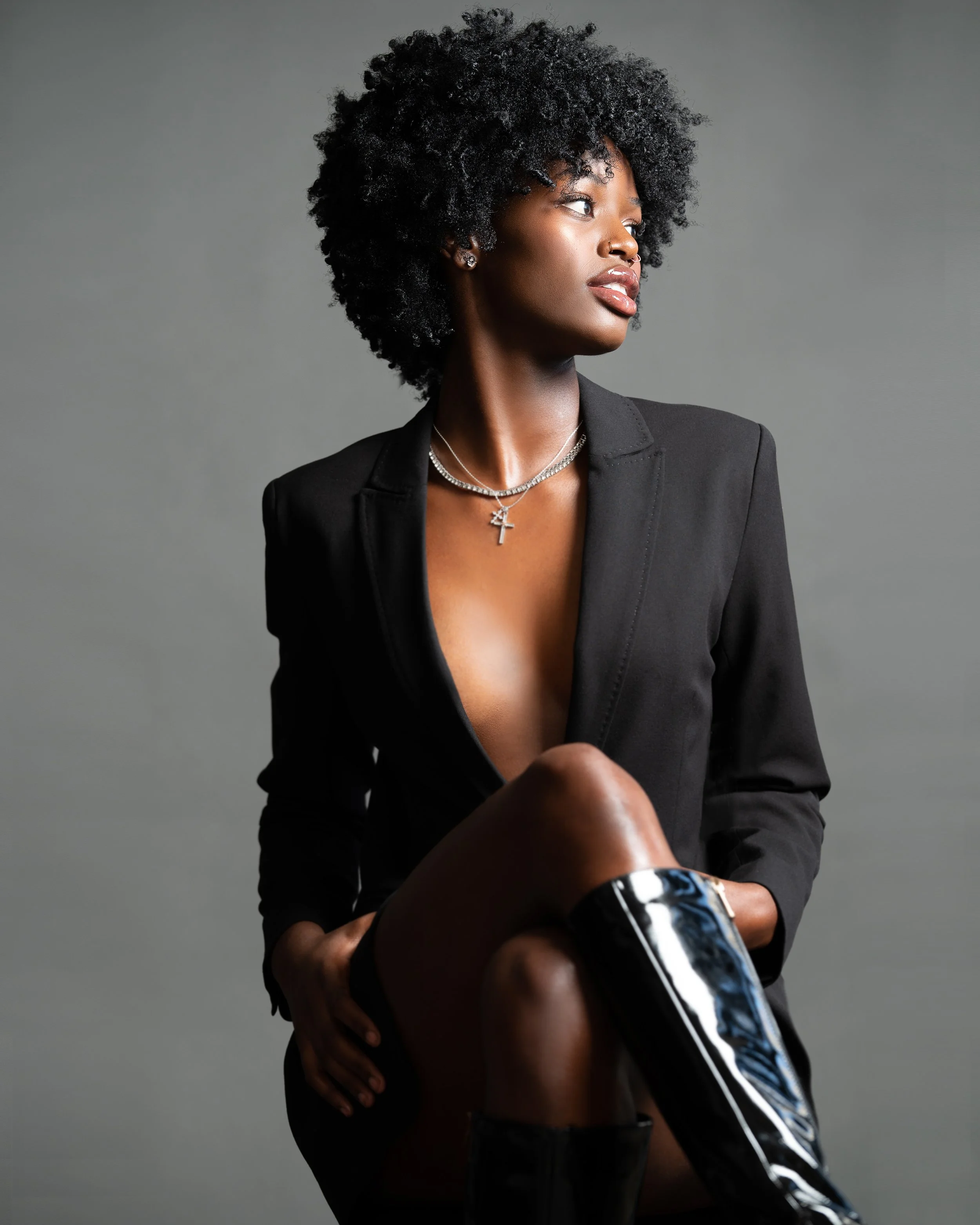 A woman with dark, curly hair wearing a black blazer, silver jewelry including necklaces, earrings, and a nose ring, holding a bottle and sitting against a gray background.
