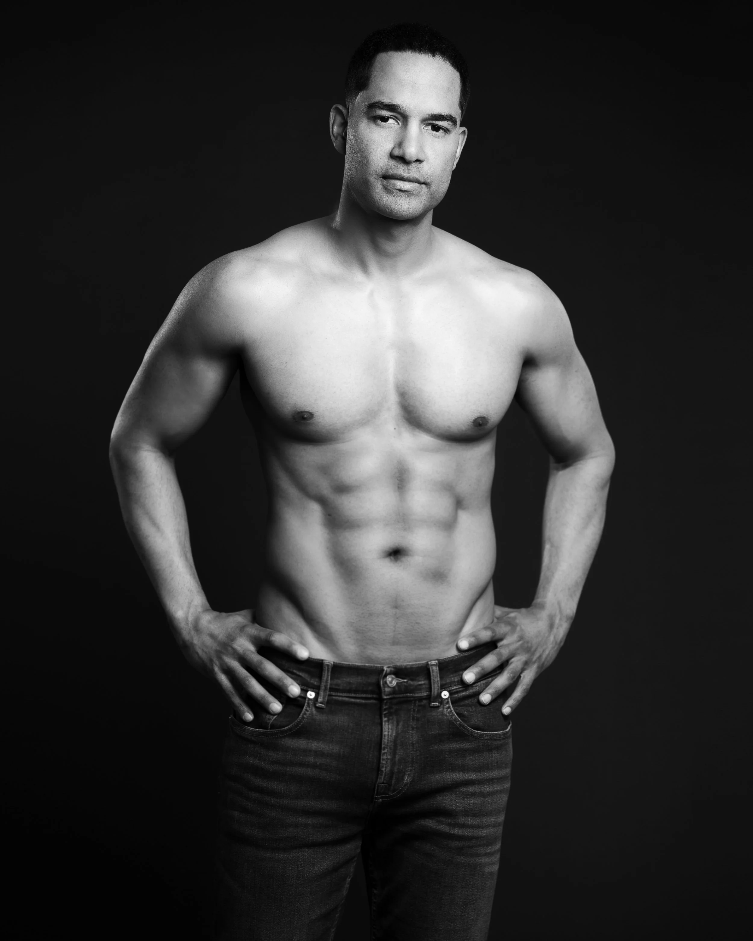 Black and white photograph of a shirtless man with a serious expression, standing with his hands on his hips against a dark background.
