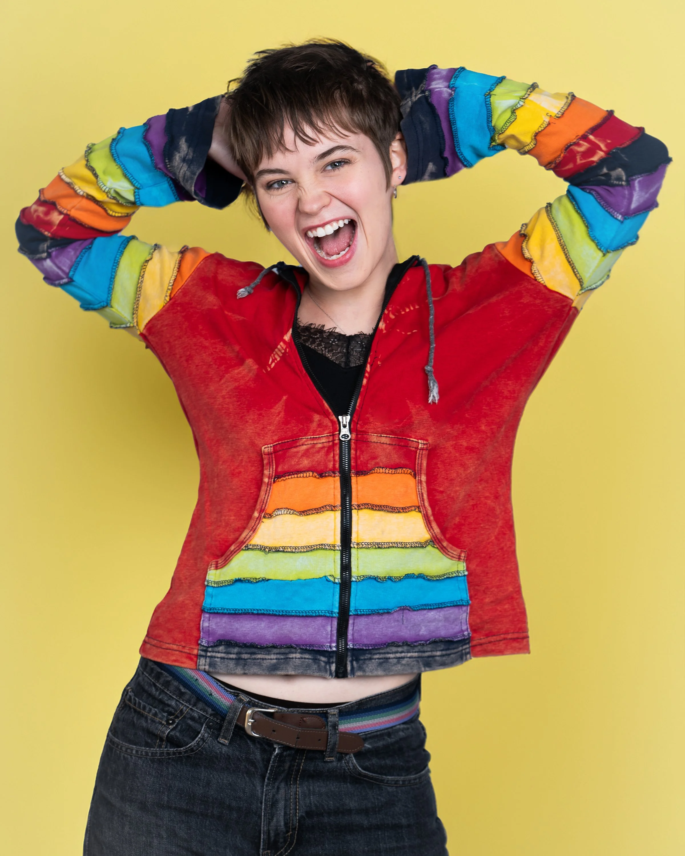 Person with short brown hair smiling and holding hands behind head, wearing a rainbow-colored tie-dye hoodie and dark jeans, standing against a yellow background.