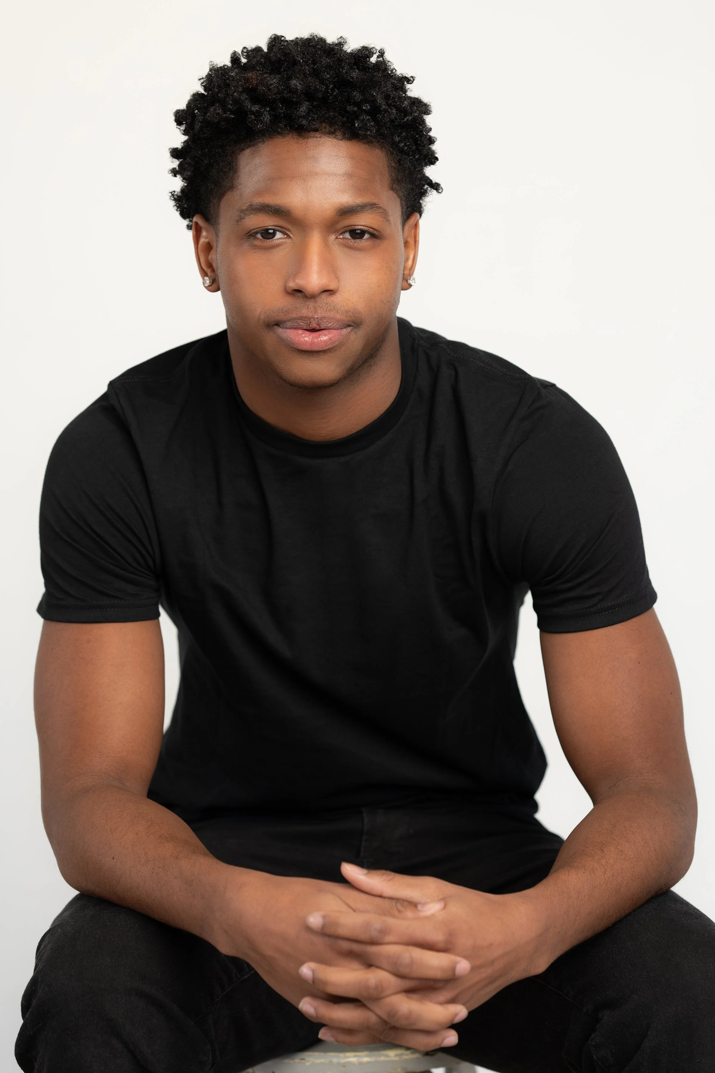 A young man sitting with his hands clasped in his lap, wearing a black t-shirt and black pants, with short curly black hair, earrings, and a light brown complexion, looking directly at the camera against a plain white background.
