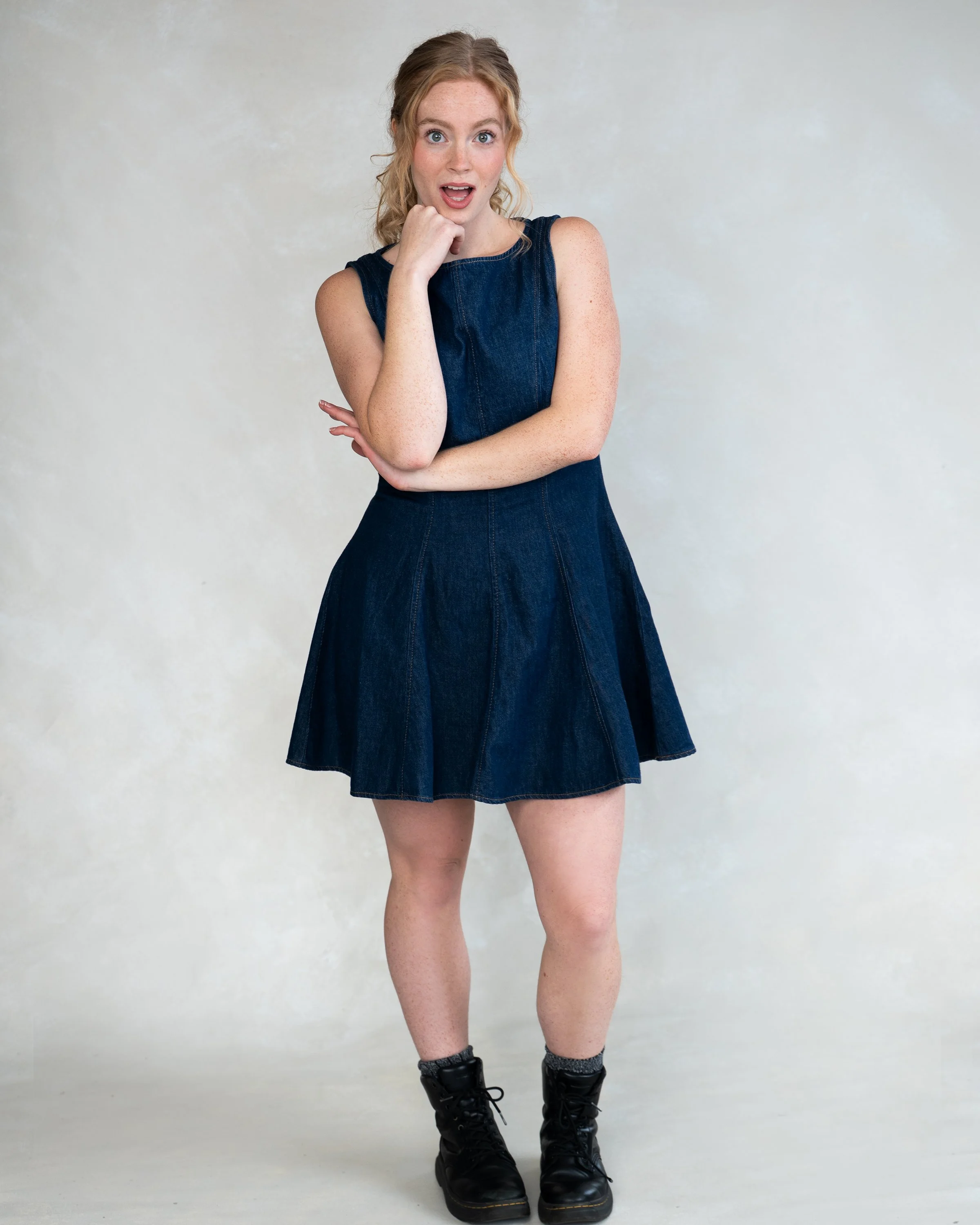 Young woman in a denim dress and combat boots standing against a plain light-colored background, with a surprised expression and hand on her chin.