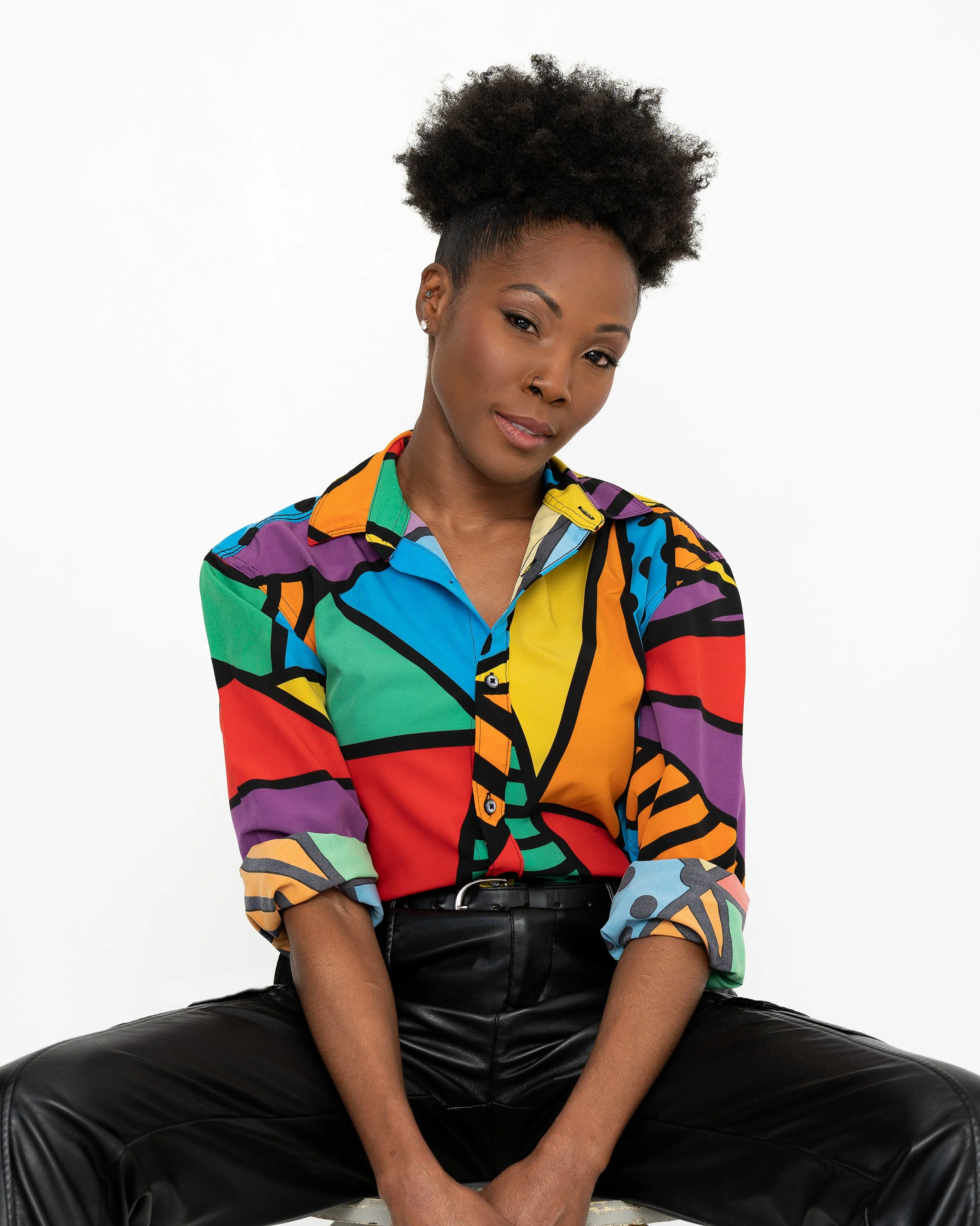 A woman with dark skin and natural hair styled in an afro is sitting in a relaxed pose, wearing a colorful, abstract-patterned shirt and black leather pants, against a plain white background.